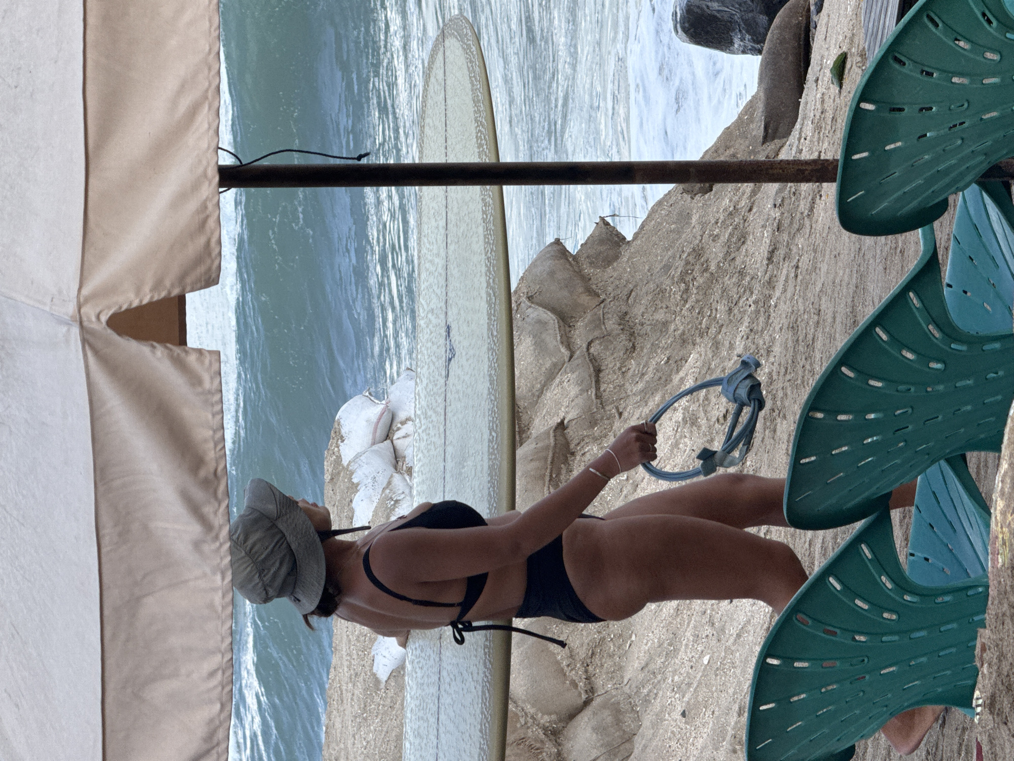 A woman wearing a dark bikini and a sun hat walks on a sandy beach near the ocean in Kuta, Indonesia. She is carrying a surfboard and a blue mask with a snorkel. In the foreground, two green beach chairs are visible. The ocean has large waves, and the sky is overcast. There are sandbags piled up behind the woman, and a seal is visible lying on the beach in the background. The scene appears to be part of a beach resort or surf spot.