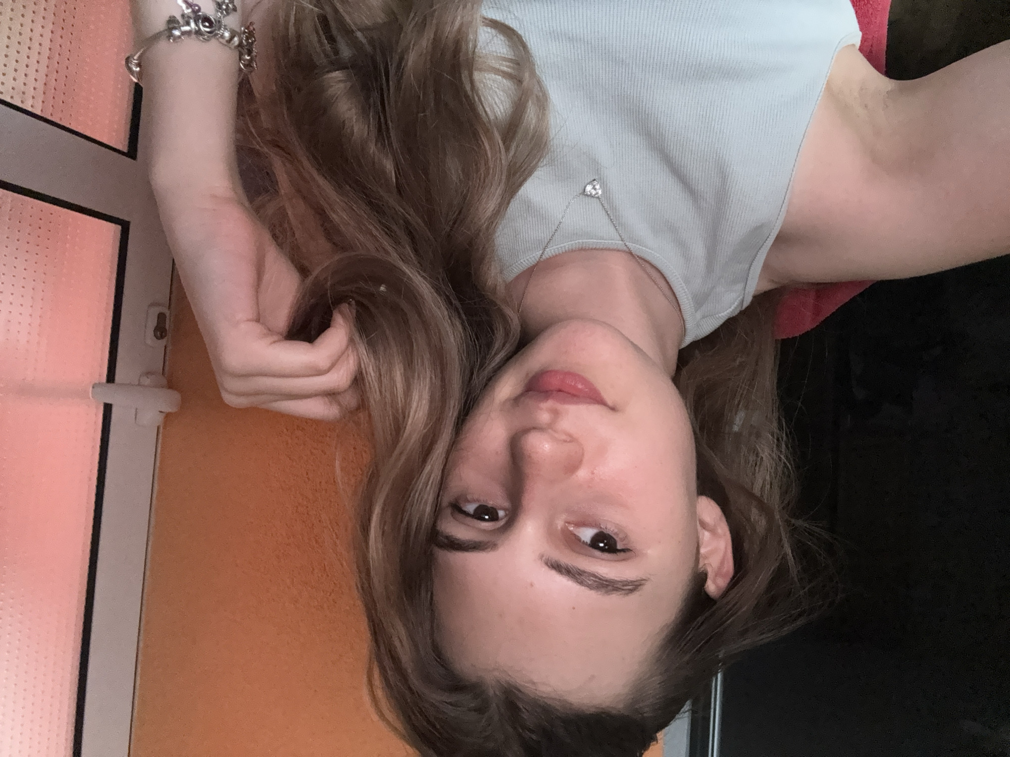 A young woman with long, wavy brown hair is shown in a close-up, frontal, selfie-style shot. She is wearing a light blue ribbed tank top and a silver necklace with a small heart-shaped pendant. Her left arm is bent, with her hand resting near her head, and her right arm is visible on the right side of the frame, adorned with a silver charm bracelet. She has fair skin, brown eyes, and is looking slightly off to her left. The background consists of an orange textured wall on the right and a dark surface on the left. A sliver of a white door frame with a handle is visible on the far right. The image does not provide any specific context that would allow for its location in Panagyurishte, Bulgaria.