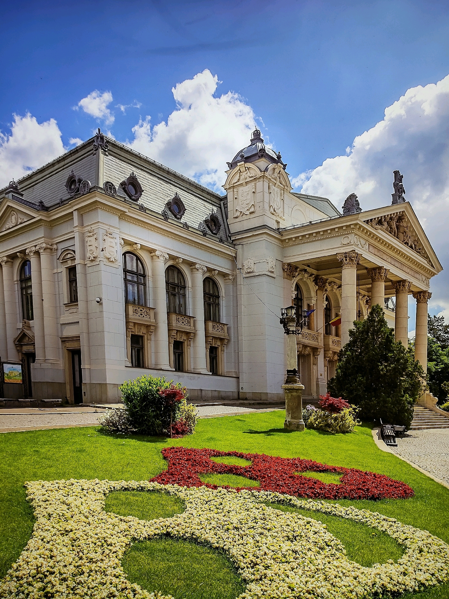 The image shows the exterior of the National Theatre of Iași, located in Iași, Romania. The building is a grand, white neoclassical structure with a prominent portico supported by Corinthian columns. Above the portico, a triangular pediment is adorned with intricate sculptures. The theater has a large, ornate roof with dormer windows and a central dome.
In front of the theater, a well-maintained lawn features a flowerbed with a striking floral design. The design consists of white and red flowers, arranged to form a decorative pattern on the green grass. The sky is a vibrant blue with fluffy white clouds. A tall lamp post stands to the left of the portico, and a dark green evergreen tree is situated to the right. In the background, a set of stone stairs leads up to an entrance, and a park bench is visible near the stairs. The overall scene conveys a sense of elegance and cultural significance.