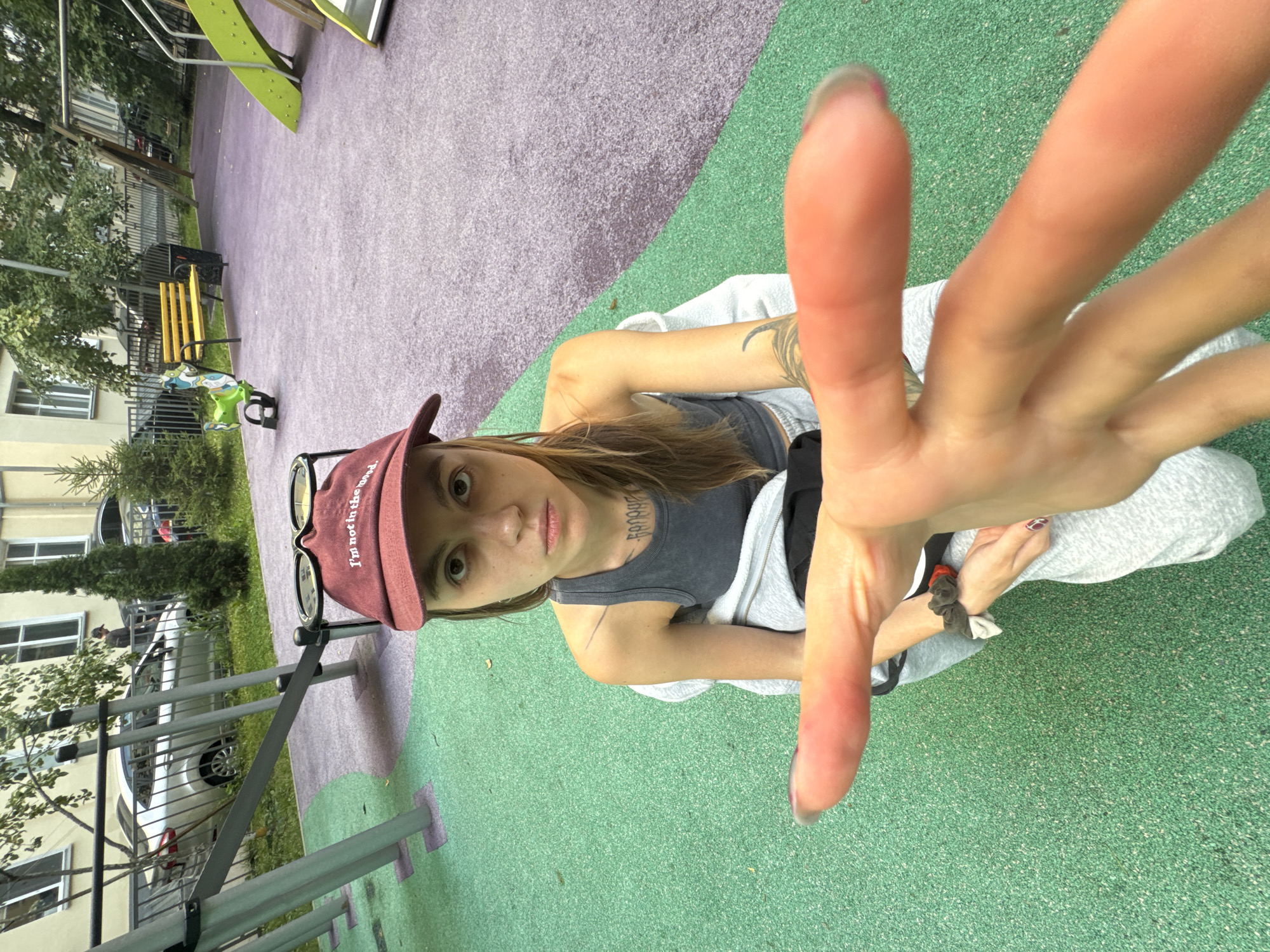 A young person is shown in a close-up selfie, taken from a low angle. They are wearing a maroon baseball cap with the text "I'm not in the mood" embroidered in white. A pair of sunglasses is resting on top of the cap. The person has brown hair and is looking directly at the camera with a neutral expression. They are wearing a dark gray tank top and a light gray hooded jacket, with grey sweatpants. A tattoo is visible on their left arm. Their right hand is prominently featured in the foreground, with fingers extended towards the camera, partially obscuring the face.
The setting appears to be an outdoor playground with a soft, multi-colored rubberized surface, featuring sections of green and purple. In the background, there is playground equipment, including a green slide. To the left, a yellow bench and a metal fence are visible, along with trees and a building with windows. A parked car can be seen behind the fence.
The image is taken during the daytime, with natural lighting. The overall mood is casual and informal. The location context is Moscow, Russia, suggested by the presence of typical apartment buildings and a playground layout common in the region.