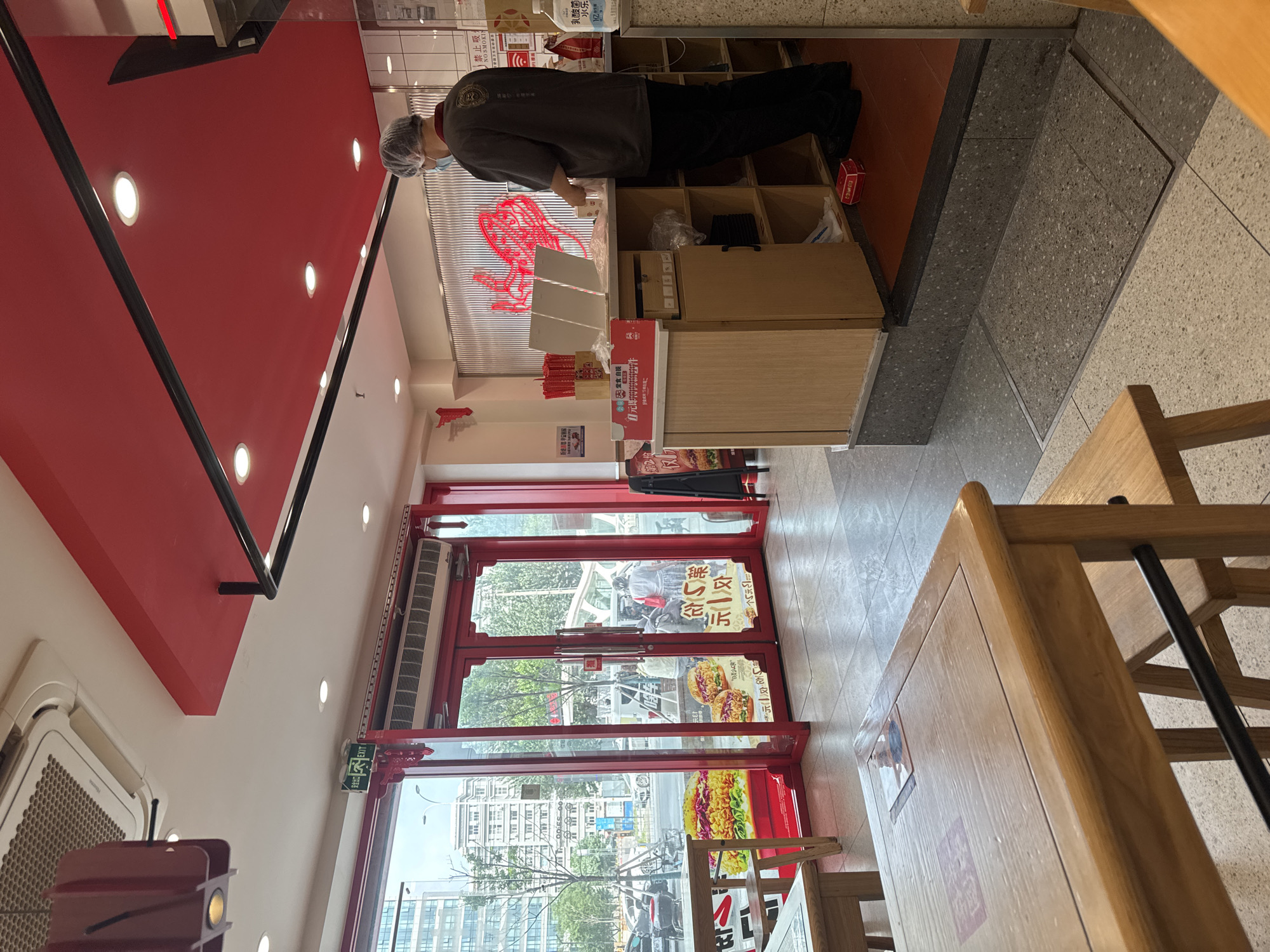 The image depicts the interior of a fast-food restaurant, likely a chain specializing in burgers or similar fare, located in Dadong District, China. The restaurant features a vibrant red and white color scheme, with red accents on the ceiling, doors, and decorative elements.
In the foreground, two wooden tables and benches are visible, suggesting a dining area. One table in the lower left has a menu or placemat on it. The flooring appears to be a light-colored tile with a grey speckled pattern.
The middle ground shows the entrance to the restaurant, with large glass doors that offer a view of the street outside. Several patrons are visible through the doors, and the street scene includes buildings, trees, and vehicles. A prominent feature on the glass doors is a large advertisement for burgers.
In the background, a service counter is set up where a restaurant employee, wearing a hairnet and a dark uniform, is interacting with what appears to be a customer. The employee is standing behind the counter, which is made of light wood and grey stone. There are shelves behind the counter with various items, including what looks like takeaway containers and bottles of hand sanitizer. A large red graphic resembling a stylized heart or hand is mounted on the wall behind the counter.
An "EXIT" sign with green lettering is visible on the ceiling near the entrance. The overall impression is that of a clean and modern fast-food establishment.