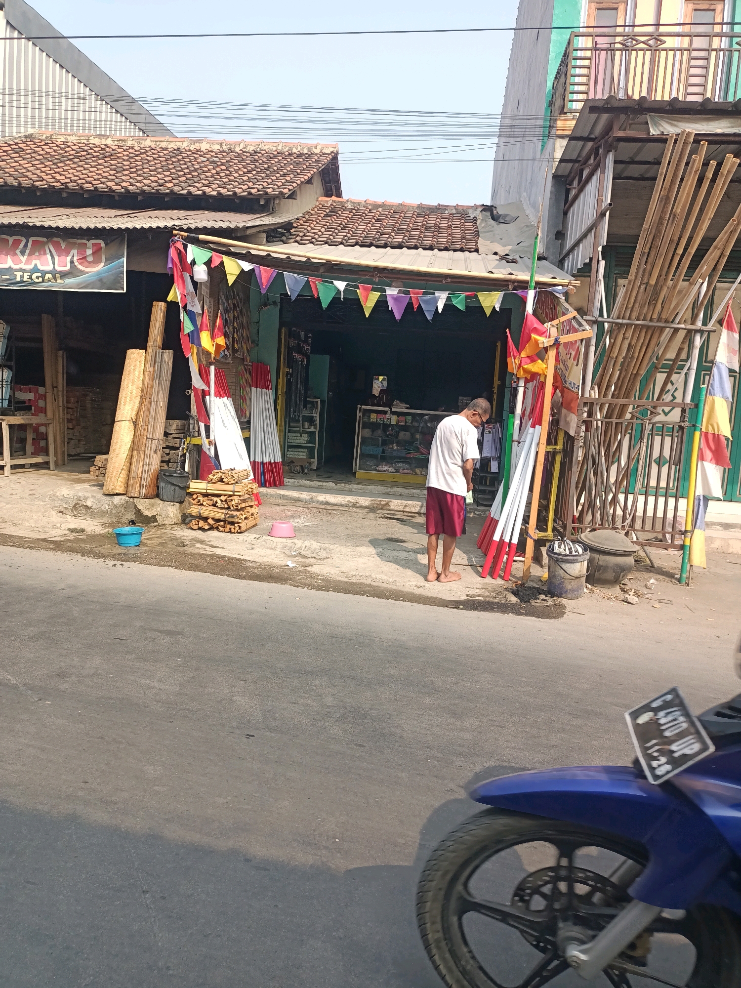 The image depicts a street scene in Tembok Banjaran, Indonesia, outside a shop that appears to sell building materials and possibly local crafts. A man, wearing a white t-shirt and red shorts, stands barefoot on the dusty ground in front of the shop, facing away from the camera. He is in the process of urinating.
The shop itself has a tiled roof and an open front displaying various items, including what looks like bamboo poles and bundles of wood. Colorful triangular flags are strung across the shop's entrance. Various stacks of wood and other materials are piled outside the shop.
To the right of the shop, a fence made of vertical bamboo poles encloses an area with more bamboo. A dark-colored bucket and a larger, dark pot are situated near the fence.
In the foreground, the asphalt road is visible, with a shadow cast across it from the left. The front of a blue motorcycle is also in the lower right corner of the image, its license plate reading "G 4970 UP" with a date of "11-26" visible. The overall environment suggests a sunny day in a somewhat urban or semi-urban setting in Indonesia.
