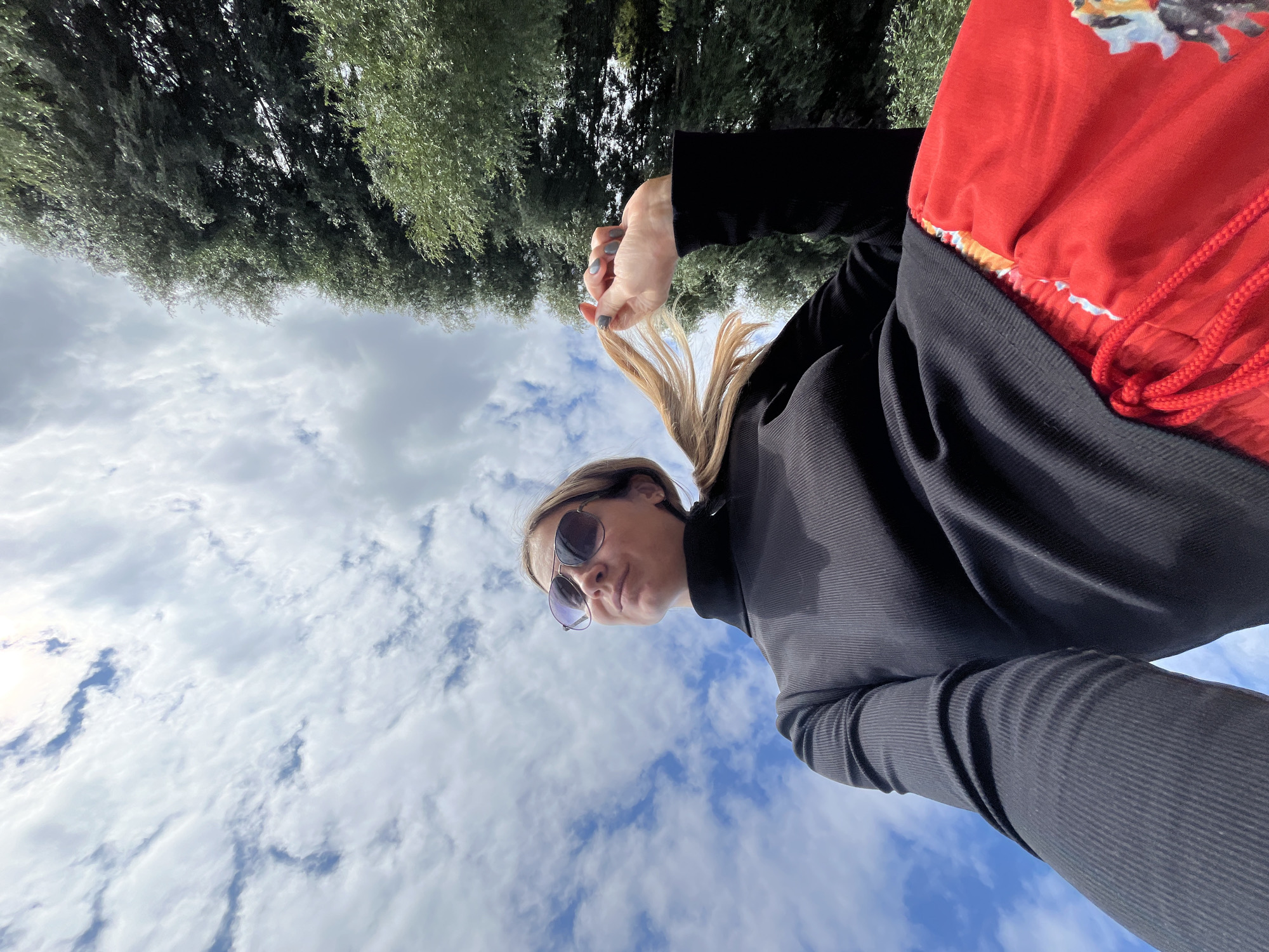 A low-angle, full-body shot captures a woman standing outdoors with a cloudy sky and trees in the background. The woman, positioned in the lower half of the frame, wears a black turtleneck and bright red patterned pants. She is looking up and to her left, her right hand is lifting a strand of her blonde hair. Her hair is pulled back into a ponytail, and she wears aviator sunglasses. The scene appears to be in Chaikovsky, Russia, given the context provided.