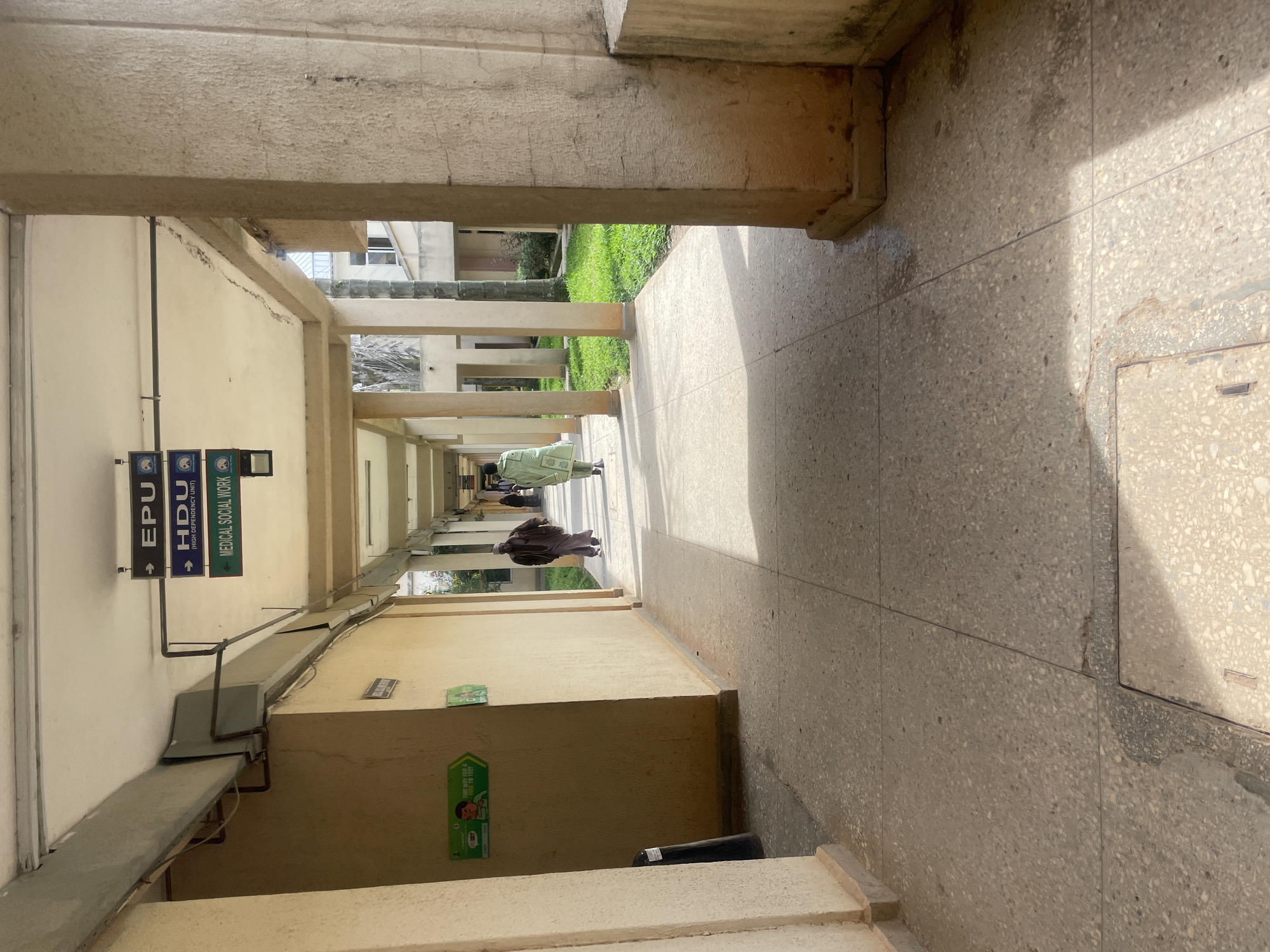 The image depicts an outdoor hospital hallway in Gwong, Nigeria. The hallway is covered by a roof supported by concrete pillars. Signs indicating "EPU," "HDU (HIGH DEPENDENCY UNIT)," and "MEDICAL SOCIAL WORK" are visible overhead, suggesting a medical facility. Two individuals are walking away from the camera down the hallway. One person wears a brown garment, and the other wears a light green garment. On the left side of the hallway, a yellow wall features a green sign with a depiction of a person and text reading "THIS WAY FOR A FREE TB TEST." The ground is paved with speckled tiles, and grass is visible to the right of the walkway. A metal vent or pipe system runs along the underside of the hallway roof. A black cylindrical object, possibly a gas cylinder, stands near the wall on the left. The scene appears to be during daylight, with sunlight casting shadows on the ground.