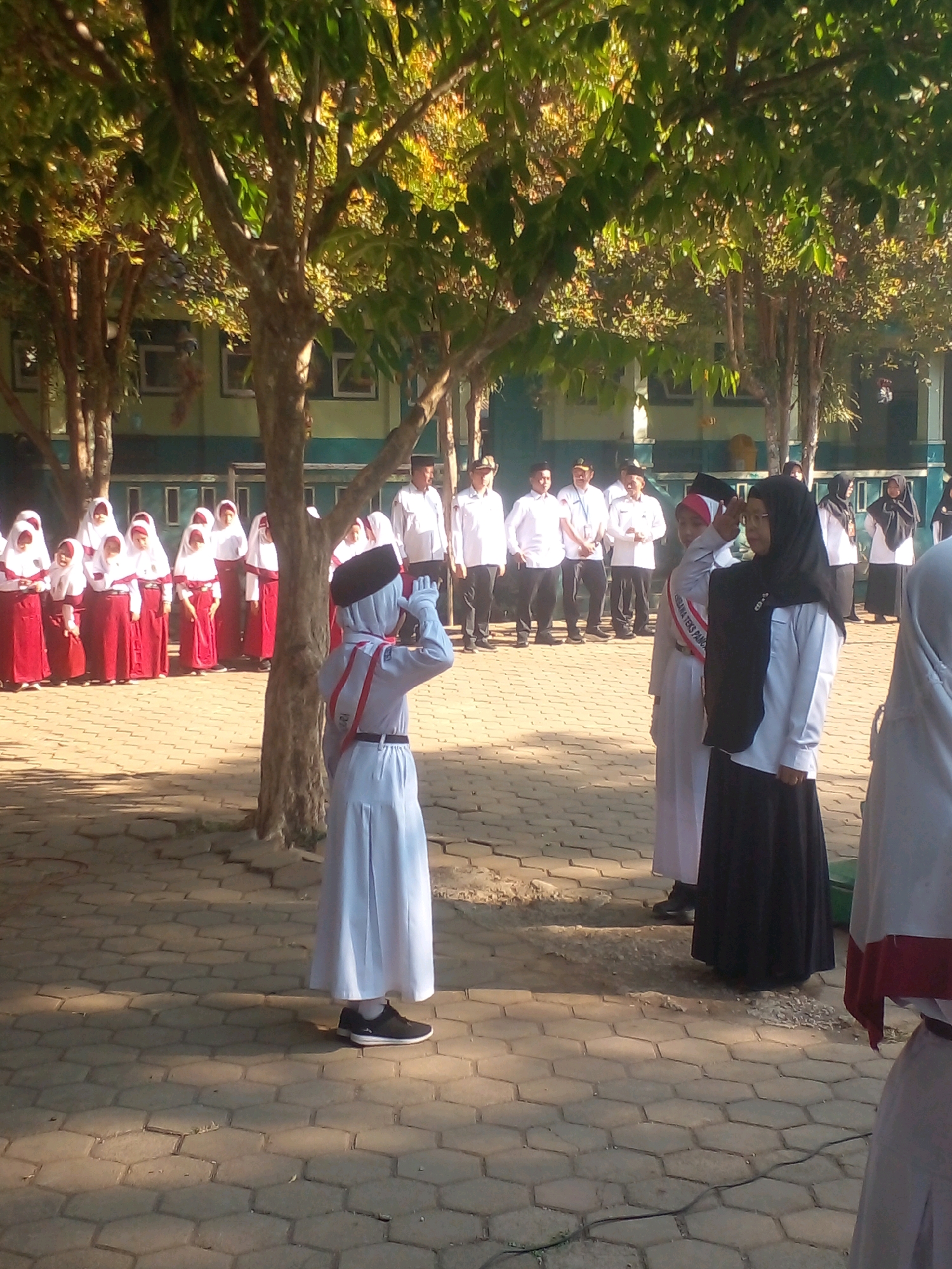 In Banyuputih, Indonesia, a group of students are participating in an outdoor event on a paved area under trees. The students, mostly girls wearing white shirts, red skirts or maroon skirts, and headscarves, are standing in formation. One student in the foreground, wearing a white uniform and a black headscarf, is saluting. Another student in a white uniform and a black headscarf is also saluting, and a third student in a similar uniform is standing beside her. In the background, several adults in white shirts and dark pants are observing. The environment features trees with green leaves and a building with green and blue accents. The lighting suggests it is daytime, with sunlight casting shadows.