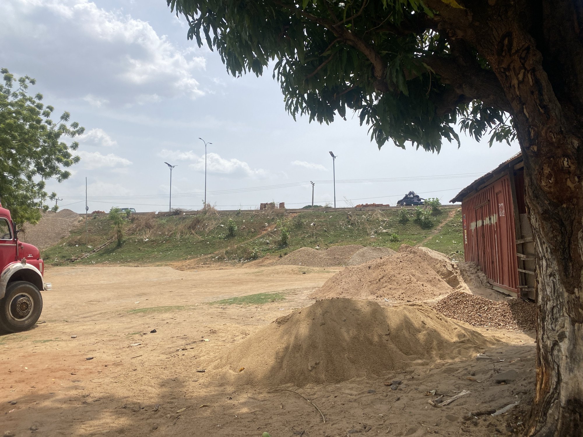 This image depicts an outdoor scene, likely a storage or undeveloped area in Makurdi, Nigeria, under an overcast but bright daytime sky. The overall ground is a reddish-brown dirt and sand, indicating a dry or arid environment.
In the foreground, a large, leafy tree, possibly a mango tree, frames the right side and top of the image, with its thick trunk and dark green foliage casting shadows on the ground. Next to the tree, a red corrugated metal structure, resembling a shipping container, is visible. Several mounds of light brown sand and smaller piles of gravel are scattered across the mid-ground.
To the far left, the front section of an older model red truck, including its large wheel and part of the cabin, is visible. The central area of the foreground consists of an open, flat expanse of dirt.
In the background, the terrain rises to an embankment covered with sparse green vegetation and some debris. Along the top of this embankment, several modern streetlights, equipped with solar panels, stand tall. Faint electrical or communication lines can be seen horizontally across the sky. Small, indistinct vehicles are barely visible on the raised area beyond the embankment.
The sky is largely covered with white and grey clouds, suggesting an overcast day, but the overall lighting is bright, consistent with late morning or early afternoon. No people are present, and no specific activities are taking place, creating a static and quiet impression. There is no discernible text visible in the image.