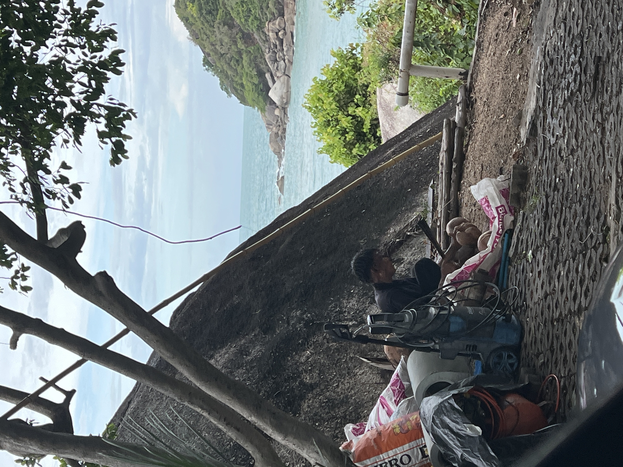 The image shows a scene in Baan Chaweng Noi, Thailand. A man is working or resting amidst a collection of equipment and belongings. The scene includes a blue air compressor, bags of various types (possibly including one with the word "ERO" visible), other tools and materials wrapped in plastic sheeting, and assorted containers. The man is located on a rocky surface near some trees and greenery. In the background, there is a view of the sea and a rocky coastline covered with vegetation.