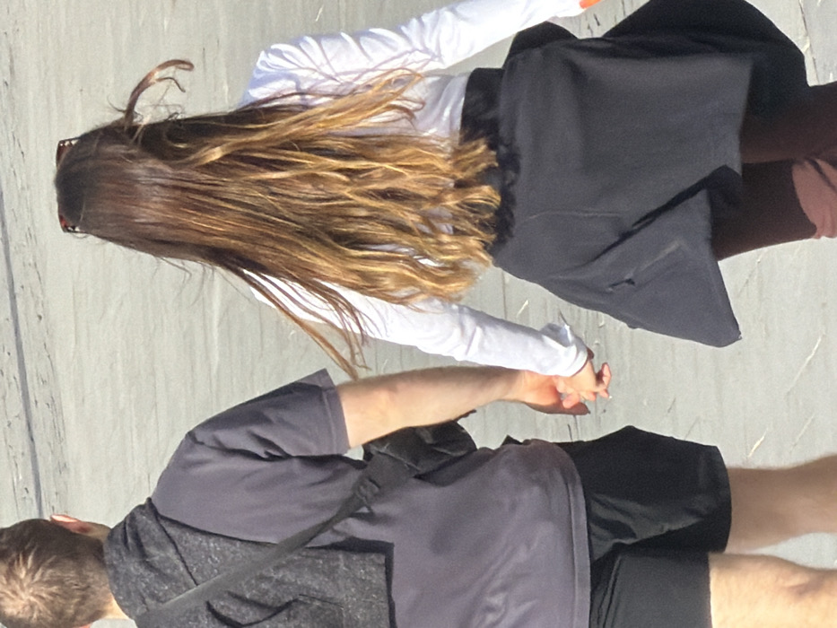 Two individuals, a male and a female, are depicted from behind walking away from the viewer in an outdoor paved area, likely a pedestrian zone, plaza, or promenade, in the city of Tel-Aviv, Israel. The specific location within Tel-Aviv is not discernible from the image.
The male, on the left, has short brown hair and is wearing a dark gray short-sleeved t-shirt and black shorts. A dark gray garment, possibly a hoodie, is draped over his left shoulder, with a black strap extending diagonally across his back, suggesting a bag or camera strap.
The female, on the right, has long, wavy brown hair with lighter, possibly sun-bleached or highlighted, ends. She wears a white long-sleeved shirt, and a dark jacket or sweater is tied around her waist. Sunglasses with tortoise-shell frames are visible resting on top of her head. Her lower garments appear to be dark brown or maroon pants.
The two individuals are holding hands, with their right hands clasped. The ground surface is light-colored and appears to be made of paving stones or concrete. The scene is brightly lit by natural sunlight, suggesting a clear day. No other individuals or distinguishing objects are clearly visible in the immediate background or foreground.