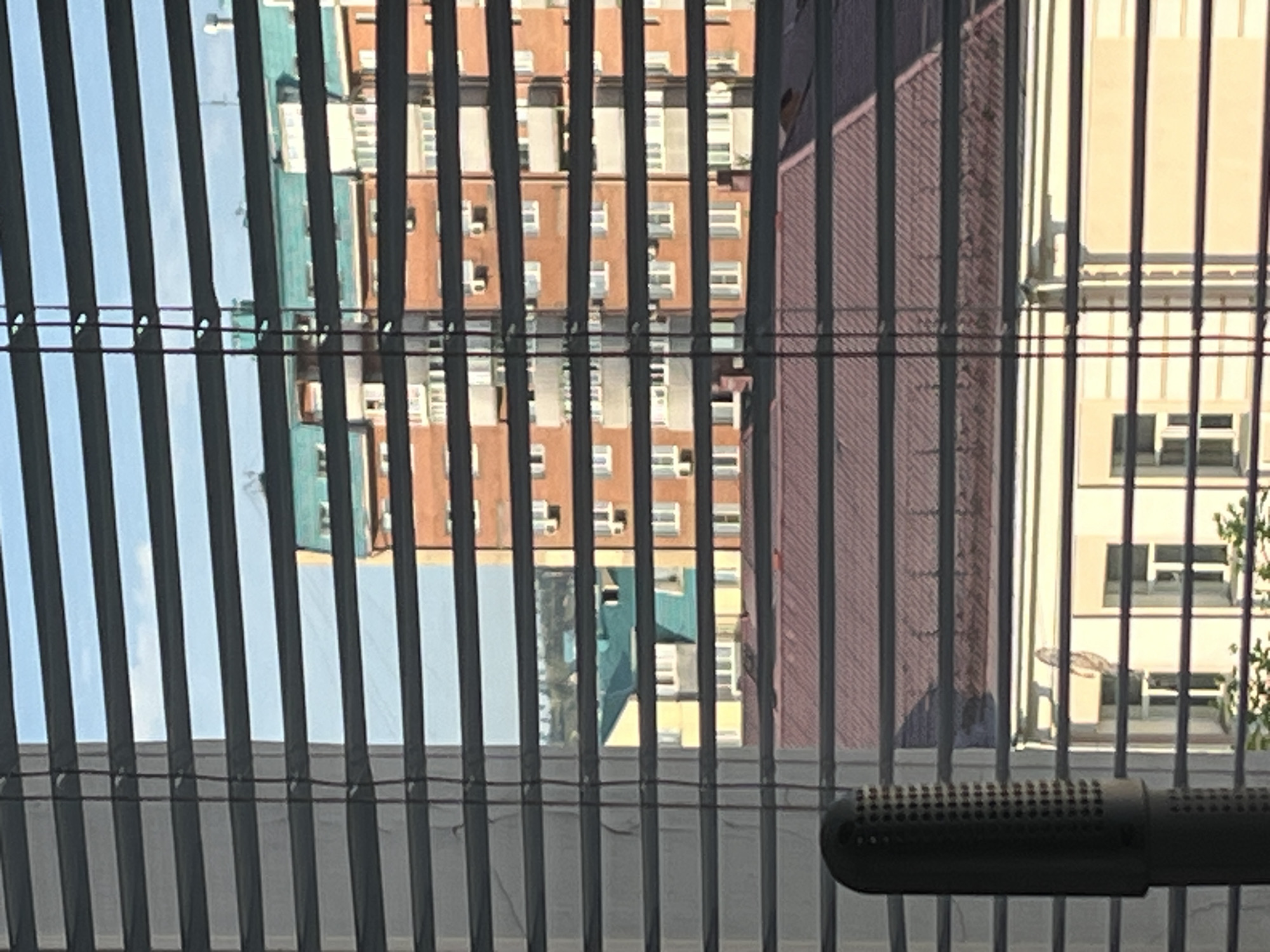 A partially obscured urban cityscape is visible through a set of horizontal blinds. In the foreground, a dark, cylindrical object with a perforated surface is present on the left side of the frame. Beyond the blinds, a large building with orange brickwork and multiple balconies and windows is prominent. To the left of this building, another structure with a teal roof is partially visible. In the foreground, a building with a reddish-brown, tiled roof is apparent, below which another light-colored building with windows can be seen. The sky is a pale blue.