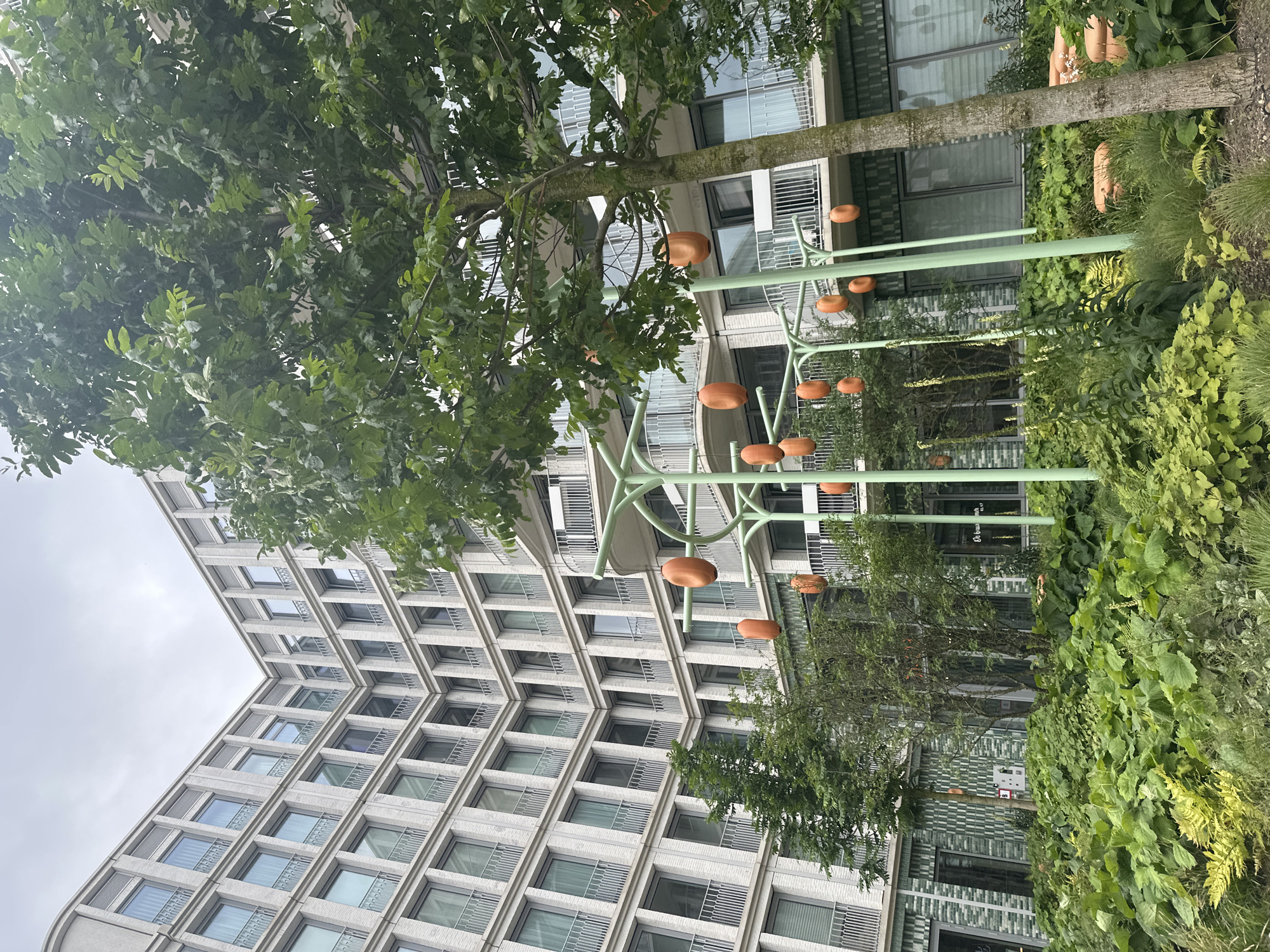 The image depicts a modern building with a curved facade constructed with light-colored bricks and numerous windows with balconies. In the foreground, there is a lush green garden with dense foliage. Several decorative elements resembling large orange pods are suspended from light green, tree-like structures. A mature tree with a textured trunk stands to the right of the frame, its branches laden with green leaves. The scene is set against an overcast sky. The building appears to be an apartment complex or office building, likely in a contemporary urban development. Research indicates this scene is likely located in Amsterdam, Netherlands, specifically at the Amsteldok building, a mixed-use development known for its distinctive architecture and integrated green spaces.