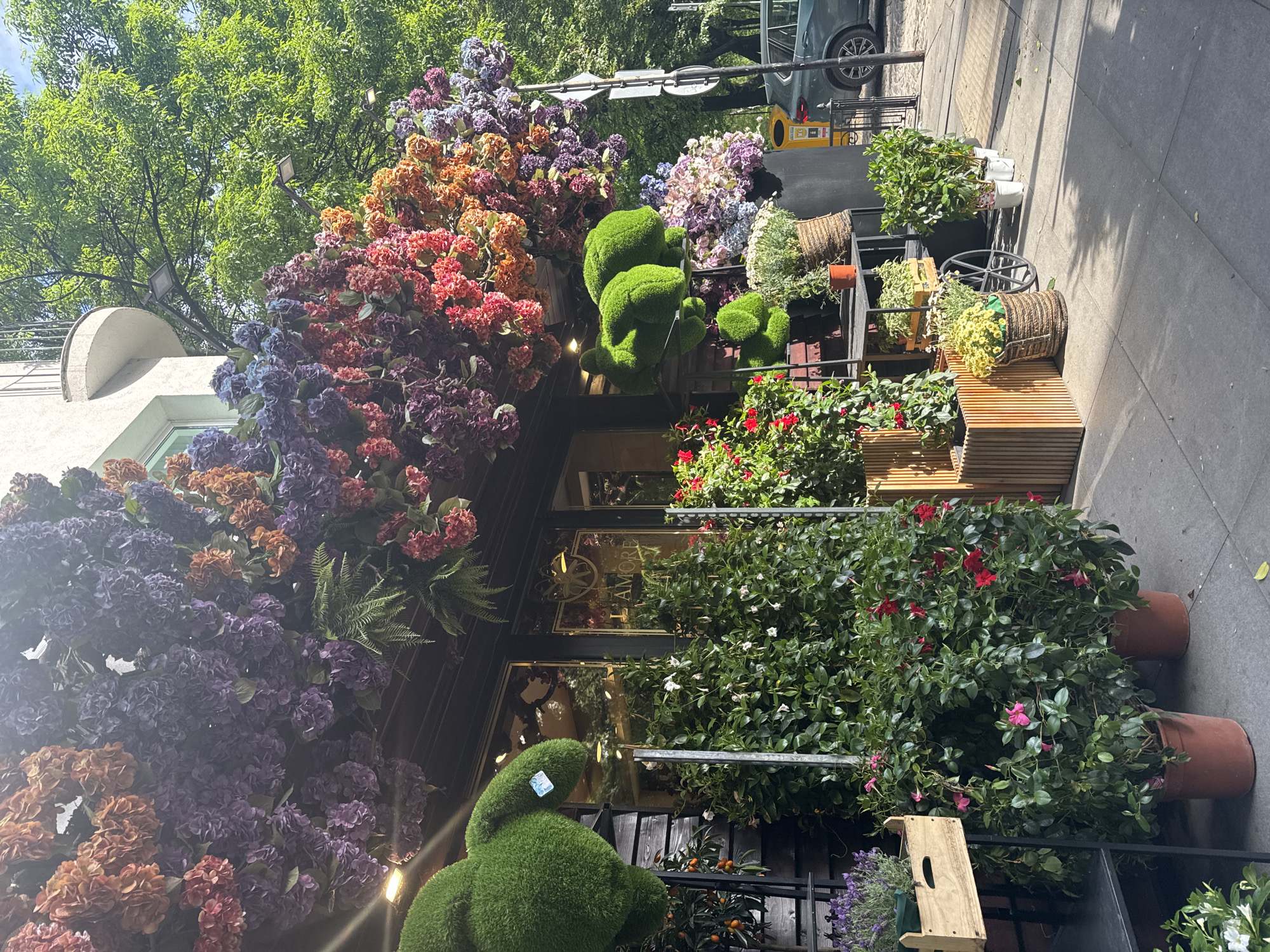 The image depicts the exterior of a flower shop in Sofia, Bulgaria, with a profusion of flowers and plants decorating the facade. The shop's entrance is adorned with a colorful array of hydrangeas in various hues, including purple, orange, and red. Green topiary figures, shaped like animals, are positioned at the shop's entrance. Potted plants, including those with red and white flowers, line the sidewalk. The shop's name, "L'AMORE," is visible on the window. A section of the sidewalk is also visible, with a glimpse of a car and traffic signs in the background. The scene suggests a vibrant and well-maintained urban environment with a strong emphasis on aesthetics.