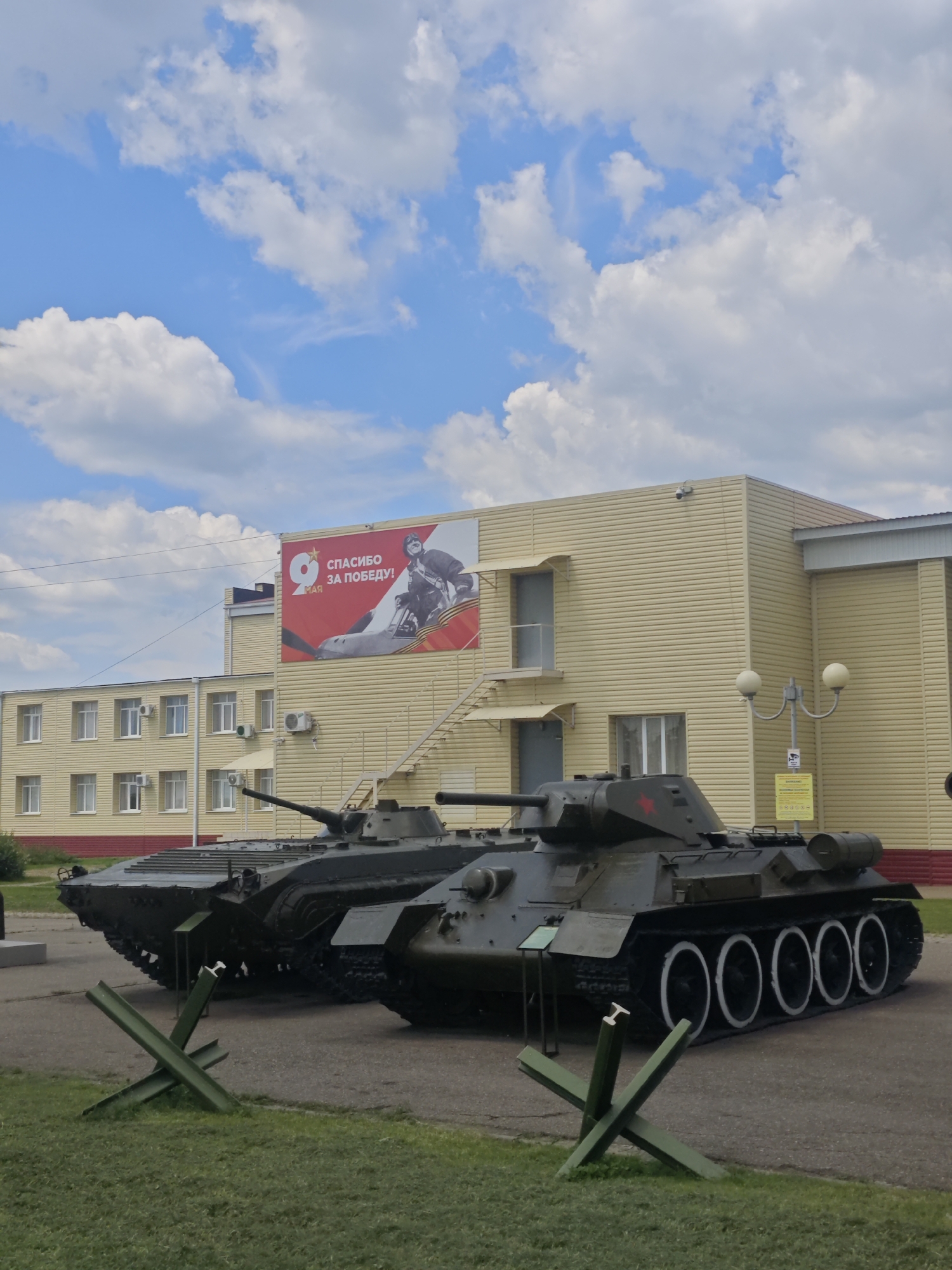 In Vyselki, Russia, an outdoor display features two military tanks positioned in front of a pale yellow building. The building has a large red banner on its facade that reads "9 МАЯ СПАСИБО ЗА ПОБЕДУ!", which translates to "May 9th Thank you for the Victory!". The sky above is blue with scattered white clouds. In the foreground, green metal anti-tank obstacles are visible on a grassy area.