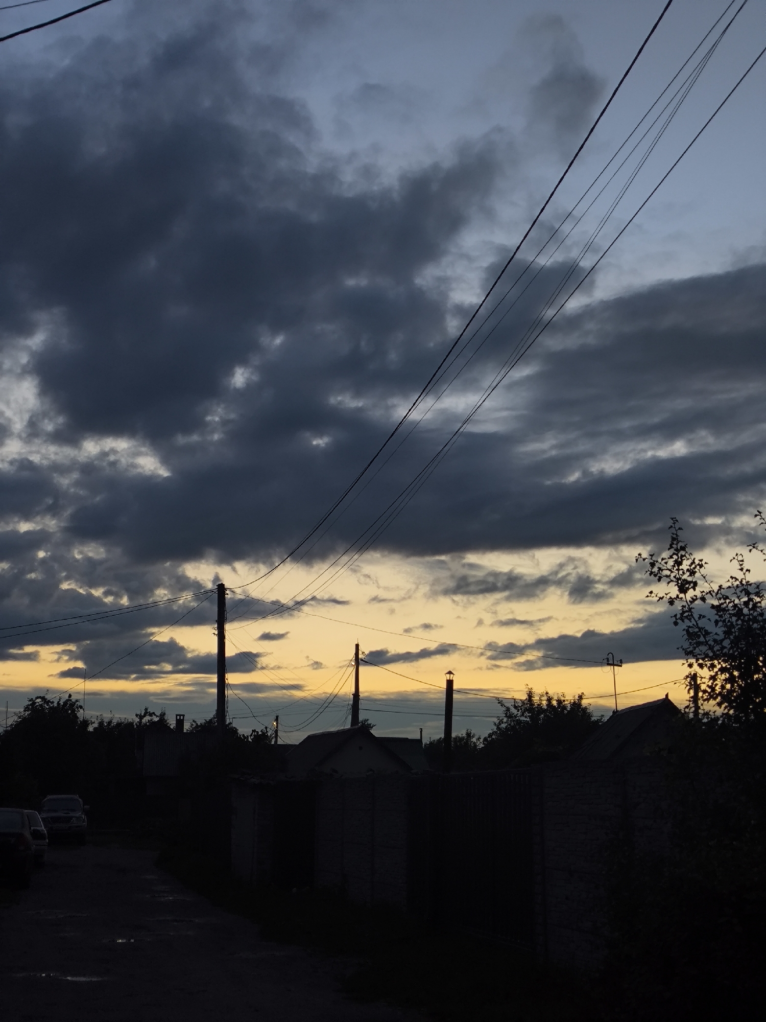 This is a medium-shot, vertical, outdoor, dimly lit photograph taken at dusk in Kaliningrad, Russia. The scene captures a rural or suburban street with houses and fences in silhouette against a dramatic sky. The sky is a gradient of deep blues and grays at the top, transitioning to a warm yellow-orange band near the horizon, indicative of sunset. Dark, heavy clouds with lighter undersides dominate the upper portion of the sky. Several power lines stretch diagonally across the frame from the top left to the top right. In the mid-ground, silhouetted against the lighter sky, are telephone poles with insulators and what appear to be chimneys or ventilation pipes atop some of the structures. Trees and vegetation are visible as dark shapes along the horizon and on the right side of the image. The foreground features a dark, unpaved road, possibly with some puddles or wet patches, and the silhouettes of two vehicles on the left. The right side of the foreground is dominated by a dark, textured fence, likely made of concrete blocks. The overall impression is one of a moody, transitional time of day in a quiet, somewhat rustic setting. There are no visible people in the image.