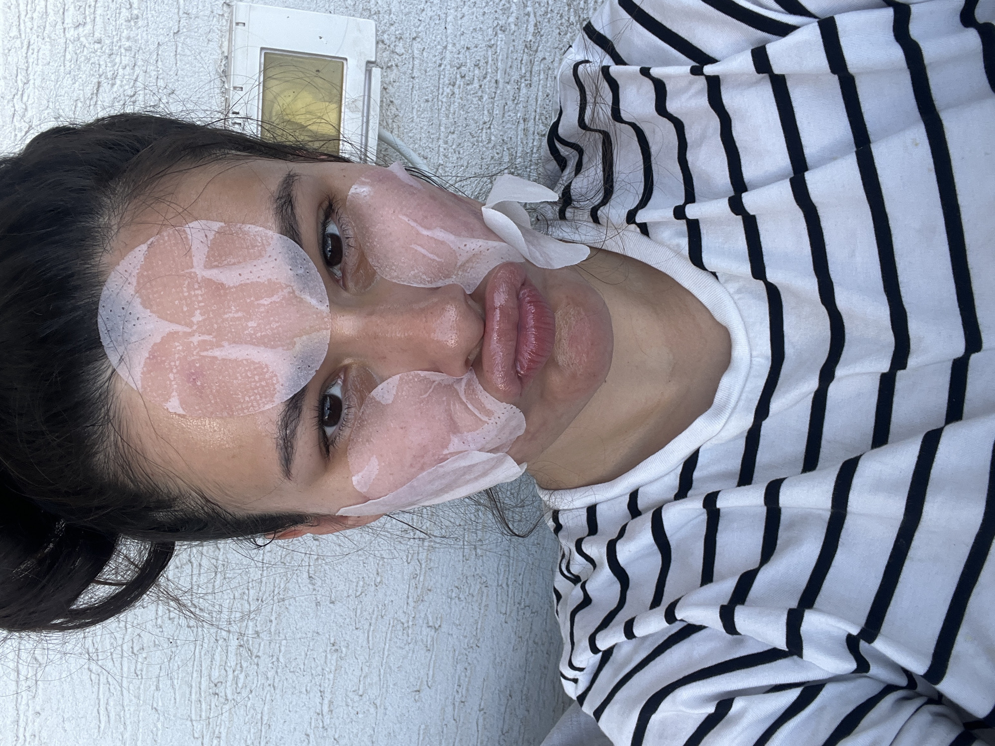 A woman is pictured indoors, wearing a black and white striped shirt and facial skincare products. These include round patches under her eyes and on her forehead and a patch over her mouth. She is lying on a light gray surface. A white electrical outlet is visible in the upper left corner of the frame. The image was taken in Kiryat Bialik, Israel.