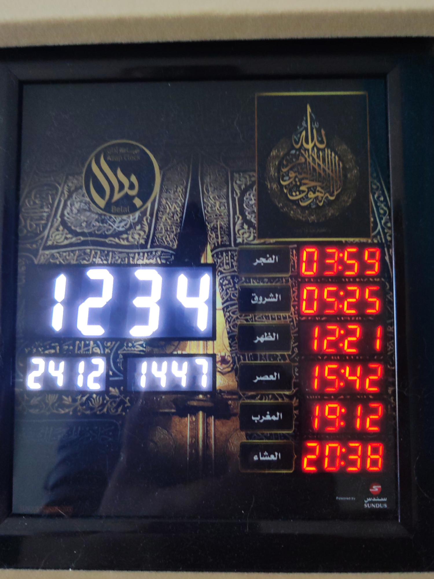 An Islamic digital clock displays the current time as 12:34 and dates 24/12 and 1447. It also shows prayer times: Fajr at 03:59, Shuruq at 05:25, Dhuhr at 12:21, Asr at 15:42, Maghrib at 19:12, and Isha at 20:38. The clock is adorned with Arabic calligraphy and an image of the Kaaba in Mecca. The clock is powered by Sundus. The overall scene is a close-up indoor shot, with the clock being the central focus. There are no people or animals present, and the background is a muted color, likely a wall or shelf. The image does not provide any information about the location being Dubai, United Arab Emirates.