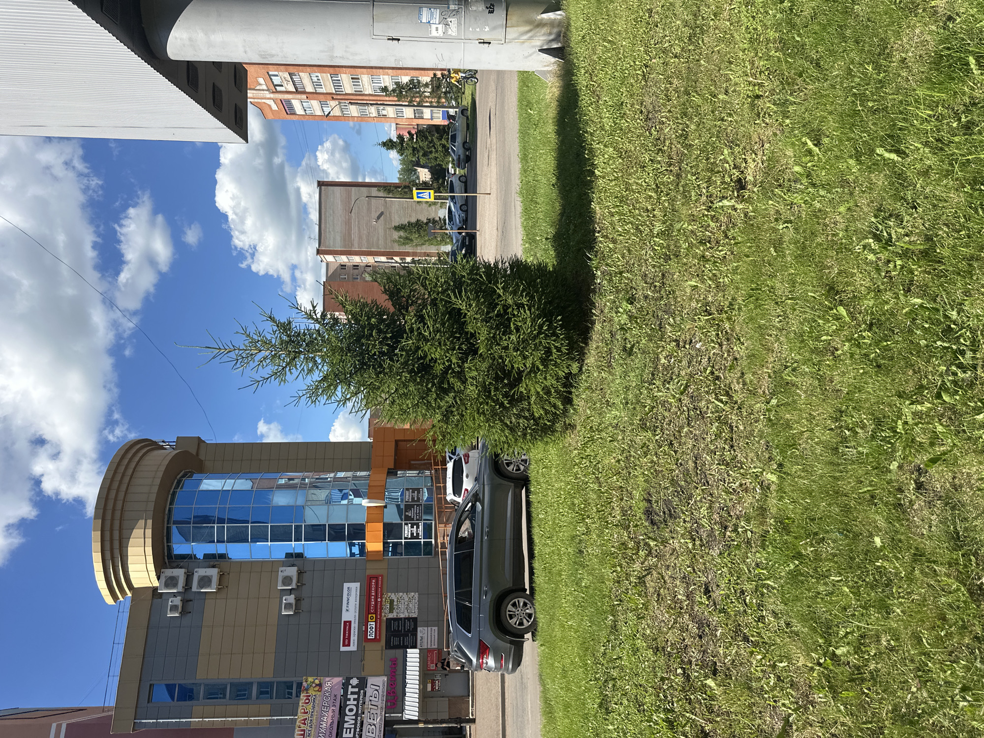 This is a photograph taken on a sunny day in Mariinsky, Russia. The image is oriented vertically. In the foreground, a grassy area with some weeds is visible. Behind the grass, a modern building with a glass facade is present, featuring blue reflective windows and several air conditioning units mounted on its exterior. Signs in Russian are affixed to the building, including "ШАРЫ ВСЕ ДЛЯ ПРАЗДНИКА" (Balloons Everything for the Holiday), "ИХМАХЕРСКАЯ" (Hairdresser's), and "EMOHT" (Repair). A gray SUV is parked on the road next to the building, and another white car is partially visible behind it. A small evergreen tree stands in front of the building. In the background, residential buildings and a pedestrian crossing sign are visible. The sky is blue with fluffy white clouds.