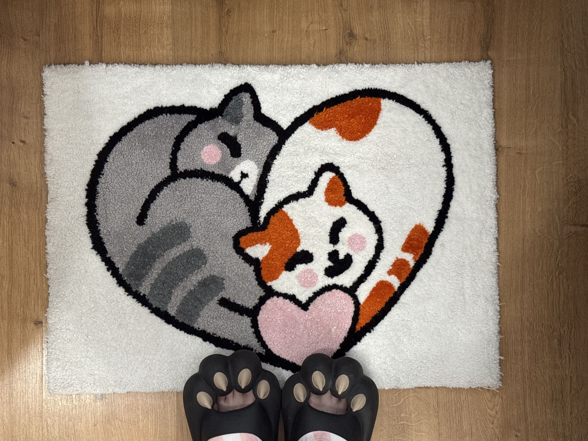 The image shows a rectangular rug with a design of two cats forming a heart shape. One cat is gray with dark gray stripes, and the other is white with orange spots. Both cats have pink cheeks and black facial features. Between them is a pink heart. Below the rug, two feet wearing black cat-paw-shaped slippers are visible. The rug is placed on a wooden floor with a light brown color. The location is not identifiable from this image.