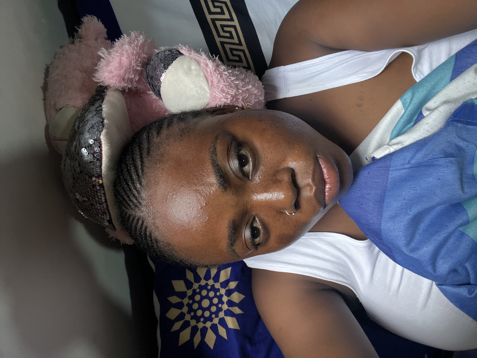 In Abuja, Nigeria, a young woman with braided hair and a nose ring lies in bed. She is wearing a white tank top and is partially covered by a blue and white patterned blanket. A pink stuffed animal with sequined ears rests behind her head. The room appears to be a bedroom with patterned pillows and bedding. The lighting suggests it may be nighttime or the room is dimly lit.
