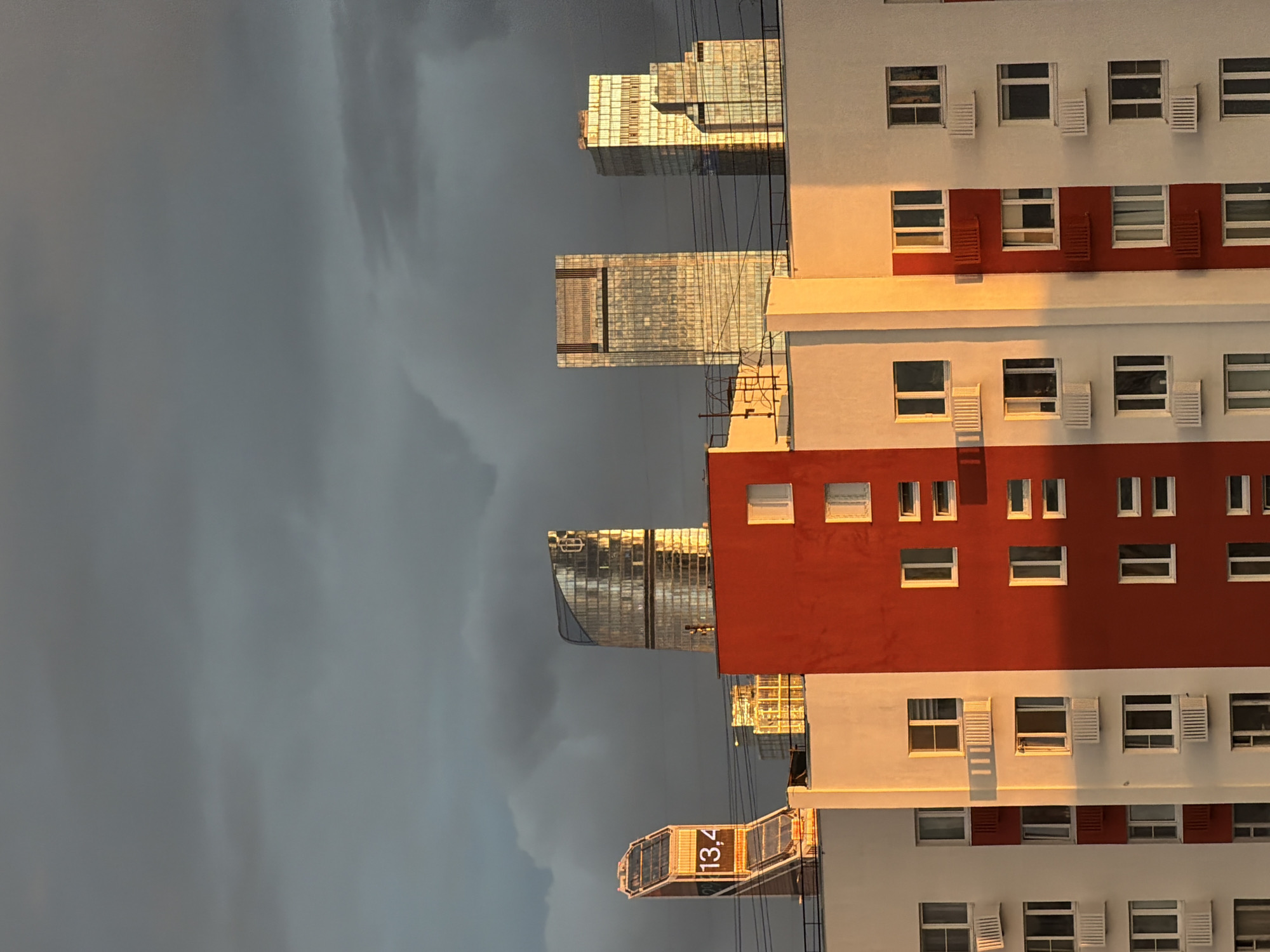 The image shows a view of buildings in Khimki, Russia, under a dramatic, cloudy sky. On the right side of the image is a multi-story building with a white and red facade, featuring numerous windows with shutters. The setting sun casts a warm glow on the building. In the background, towards the left, are modern glass skyscrapers that reflect the light. Overhead wires are visible. The sky dominates the left side of the image with dark, heavy clouds.
