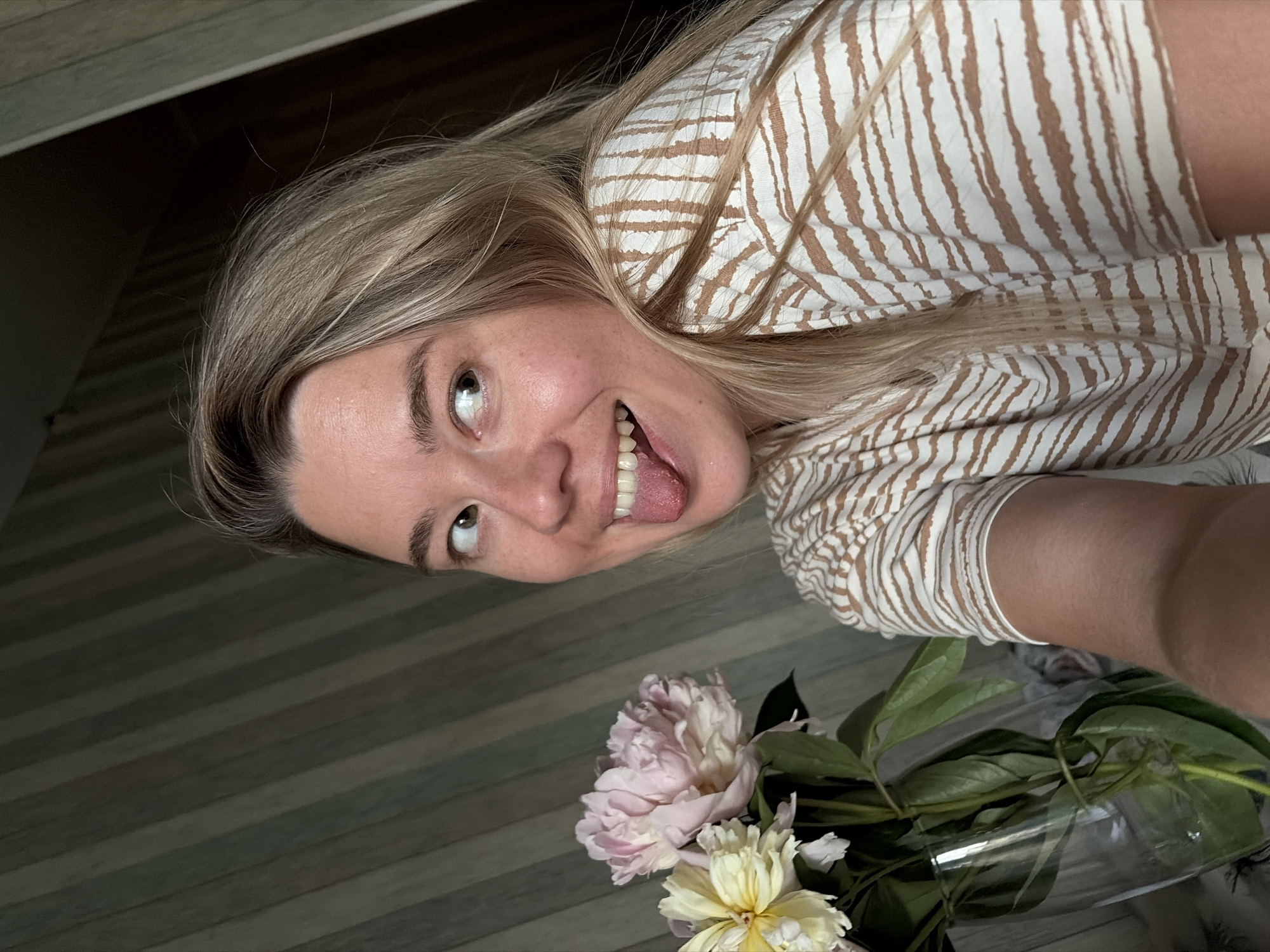 A woman is shown indoors, with a bouquet of pink and yellow peonies in a glass vase on the left. She is wearing a beige and white striped shirt and is looking to the right with her tongue sticking out. The background consists of a wall with vertical striped wallpaper. The location is not identifiable as Chaikovsky, Russia based on the visual information provided.