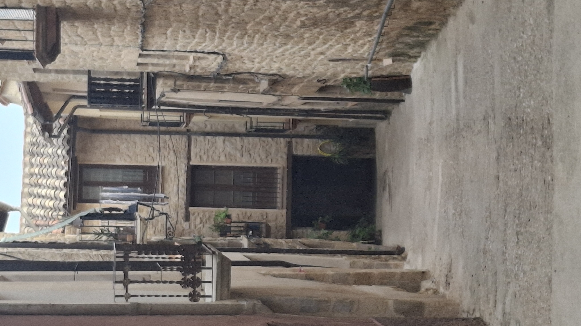 The image captures a narrow, sloped cobblestone alleyway in the historic town of La Fresneda / la Freixneda, Spain. Stone buildings line both sides of the alley, with architectural details such as balconies with wrought-iron railings and tiled roofs visible. Some windows are present, with one showing curtains. A dark, large doorway or garage entrance is situated at the end of the visible alley. Greenery in the form of potted plants and climbing vines adds touches of nature to the otherwise stone environment. The sky is partially visible at the top of the image, suggesting it is daytime. The overall impression is one of a quiet, aged street within a Spanish village.