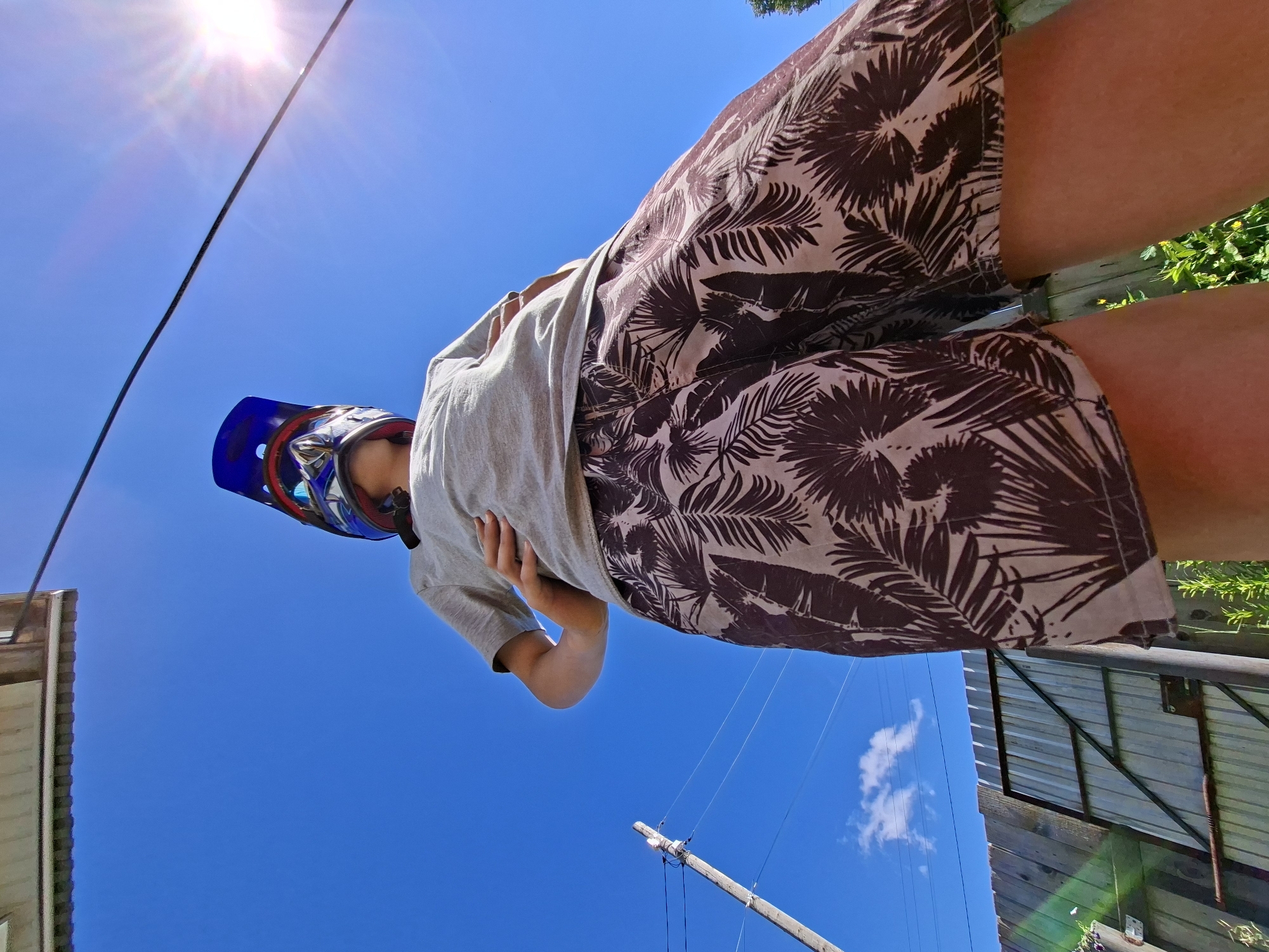 A low-angle, full-shot photograph shows a person wearing a blue motocross helmet, a gray t-shirt, and patterned shorts outdoors on a sunny day. The person stands with their left hand on their hip, facing away from the camera and looking upwards towards the sky. The background includes a clear blue sky with a bright sun and lens flare in the upper right corner, a wooden fence and gate, a utility pole with wires, and some greenery. The foreground is dominated by the person's legs and shorts.