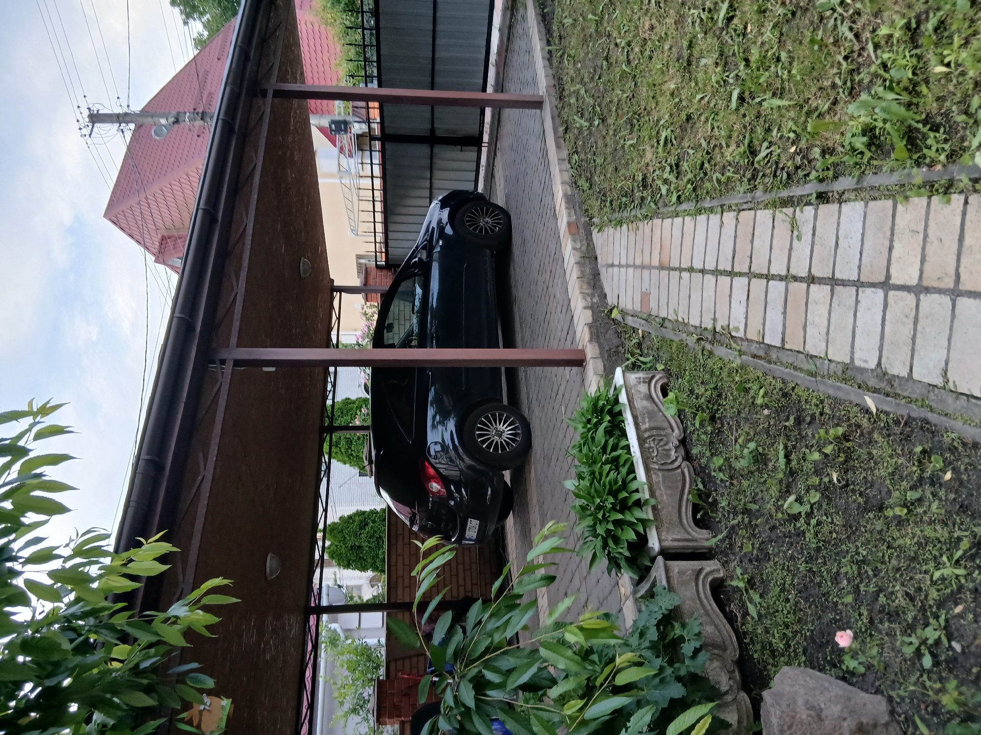 This image depicts an outdoor scene at a residential property in Vita-Poshtova, Ukraine. A black hatchback car is parked under a dark brown carport with a corrugated roof. The carport is supported by brown metal posts and sits on a paved area constructed with interlocking bricks. Adjacent to the carport is a grey metal fence. In the foreground, a path of light-colored pavers runs through a grassy area with some small plants and two decorative concrete planters. Lush green foliage from a tree overhangs the left side of the frame. In the background, a house with a red tiled roof and a utility pole with electrical wires are visible. The sky is overcast. No people are present in the image.