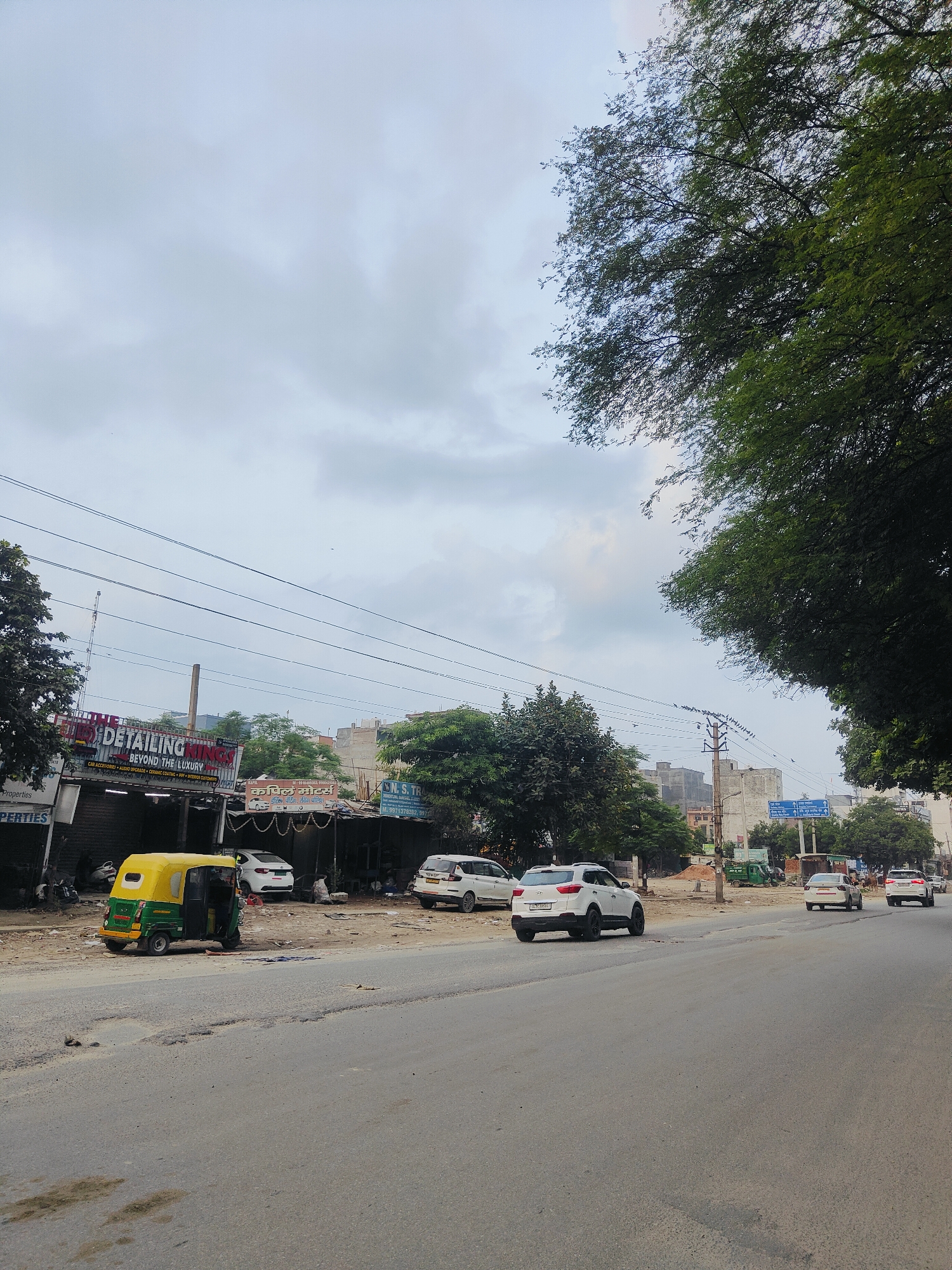 The image depicts a street scene in Gurgaon, India, on a cloudy day. A paved road with visible potholes runs through the foreground, with several vehicles present. On the right side of the road, a white SUV is driving away from the camera, followed by another white car. Further down the road, another white car is also moving away. On the left side of the road, a green and yellow auto-rickshaw is parked. Behind the auto-rickshaw, there are several small shops or businesses. One prominent sign reads "DETAILING KINGS BEYOND THE LUXURY" with smaller text indicating services like "CAR ACCESSORIES AUDIO UPGRADE CERAMIC COATING PPF UV PROTECTION". Adjacent to this is another shop with a sign in Hindi script, and further to the right, a sign for "S.N. TRADERS". Trees line the street, and buildings, including some multi-story structures, are visible in the background. Power lines crisscross the sky, supported by utility poles. The overall impression is of a commercial area in an urban setting.