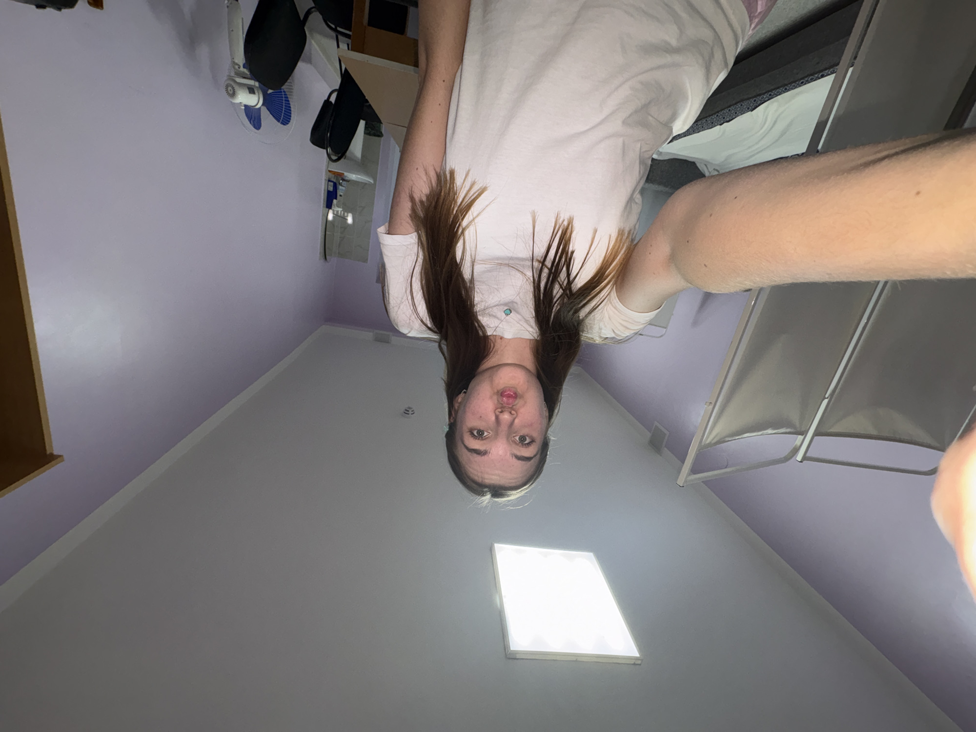 A disoriented, upside-down selfie appears to have been taken inside a room. The camera is pointed upwards towards a ceiling light fixture. A Caucasian female with long, dark hair is partially visible, her face appearing above the light. She is wearing a light-colored t-shirt. The surrounding environment includes a wall-mounted fan, shelves, a bed, and a foldable chair, suggesting a bedroom or living space. The wall color is light purple. There is no evidence to confirm that the location is Perm, Russia.