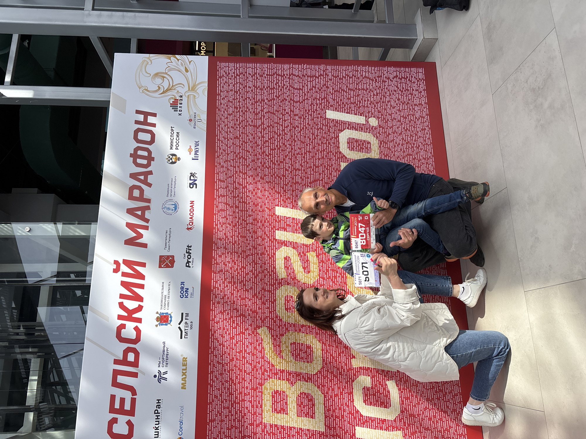 **Scene:**
The image captures a family of three posing in front of a promotional backdrop for the "Tsarskoye Selo Marathon 2026" in Saint Petersburg, Russia.
**People:**
* A woman wearing a white jacket, blue jeans, and white sneakers is kneeling on the ground, holding up a race bib with the number 5071.
* A man in a dark blue sweater and jeans is kneeling behind her, holding a young boy.
* The boy is wearing a green striped jacket and holds up another bib with the number 8047.
**Objects:**
* The main backdrop is red, filled with the names of registered participants and promotional content in gold.
* The event's official logos, including the Saint Petersburg government and sponsors, are on a poster to the left.
**Environment:**
* The location appears to be an indoor space, possibly a shopping mall or convention center.
* Reflective glass windows are visible on the left side of the image.
* The flooring is tiled with a light grey and white color scheme.