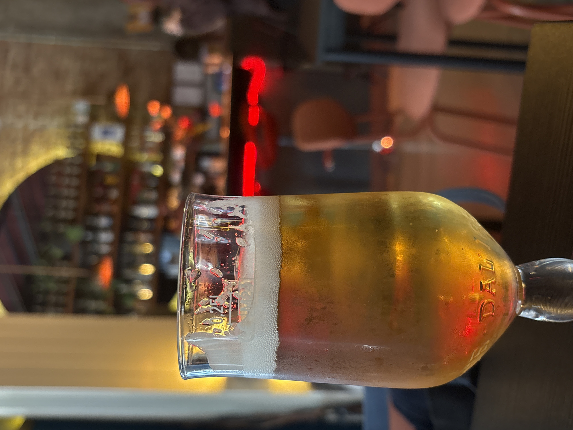 A glass of beer sits on a table, the bubbles of the foam still clinging to the glass near the top. A neon question mark hangs in the blurred background. The background also shows other bottles or glasses on shelves. The "DAL" mark on the bottom of the glass hints the location as Belgrade, Serbia. The setting seems to be a bar or restaurant.