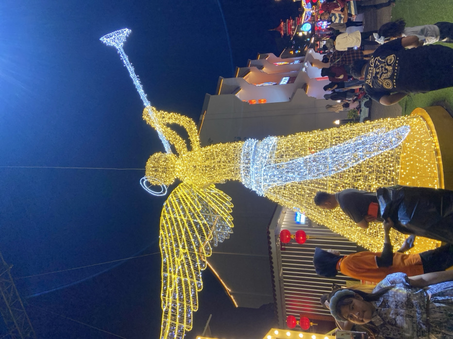 The image depicts a vibrant nighttime scene at Kya-Kya Kembang Jepun, Surabaya, Indonesia, illuminated by extensive festive decorations. A large, prominent wireframe sculpture of an angel blowing a trumpet stands centrally, constructed from myriad golden-yellow and white lights. The angel features golden wings and body, with white lights forming the trumpet, a diagonal sash across its body, and a halo.
In the foreground, a young female with light-colored hair and a light blue headband is visible on the left, wearing a patterned dress. Adjacent to the angel's base on the left, a person wearing an orange shirt and a black hijab is seen from behind, standing near a male in a grey shirt with a red armband, who has his arms crossed and is holding a black trash bag.
The background reveals architectural structures typical of East Asian design. On the right, a building with a multi-tiered roof and visible signage reading "KYA-KYA" is present. Further right, a red pagoda-like structure can be seen. On the left, another building features vertical white paneling and several suspended red spherical lanterns. Numerous other individuals, comprising adults and children, are distributed throughout the mid-ground and background, observing the decorations and moving across what appears to be a paved area combined with sections of artificial green turf. The sky is dark blue, indicating the night environment, which is brightly lit by the decorative installations.