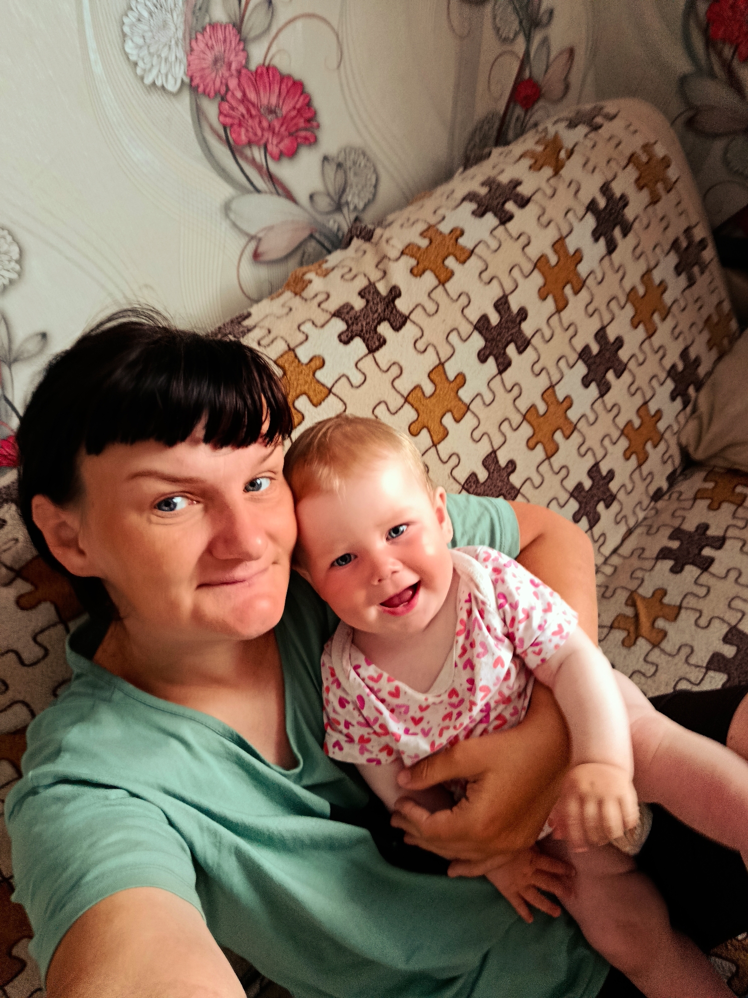 The image shows a woman and a baby indoors. The woman has short dark hair, fair skin, and is wearing a green t-shirt. She is smiling and holding the baby close. The baby has fair skin, light hair, and is wearing a white onesie with red hearts. The baby is also smiling. The background includes a wall with a floral pattern and a sofa with a puzzle-piece design. The location is Фоки, Russia.