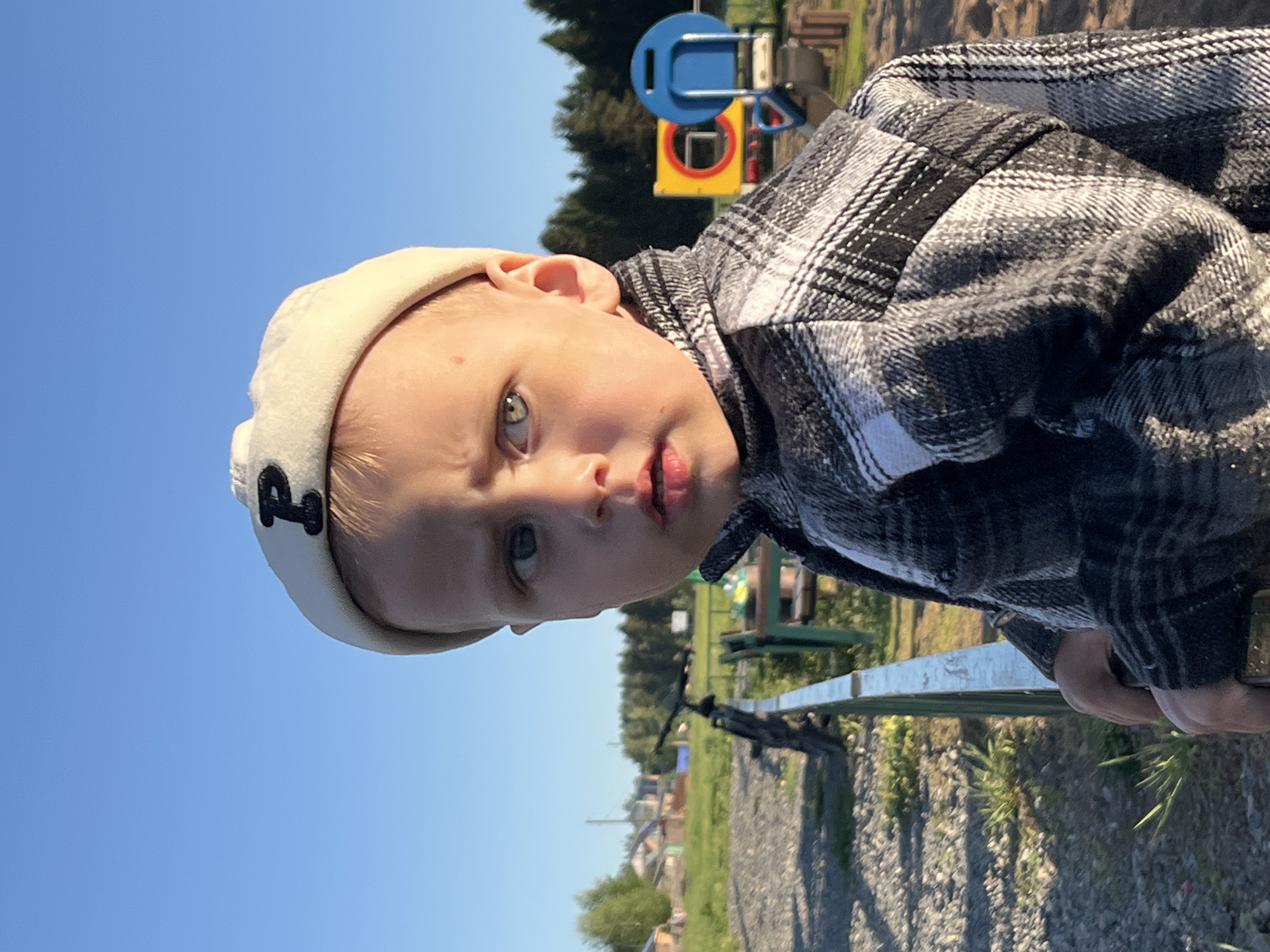 A child wearing a white cap with a black emblem and a black and white plaid jacket stands outside in a park. The background contains a green grassy area and a blue sky. In the distance, a playground with blue and yellow elements can be seen. The location is likely the city of Фоки, Russia.
