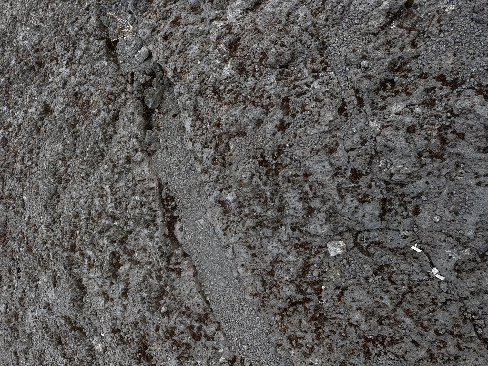 The image shows a close-up, overhead view of a rocky, uneven surface, likely volcanic rock. The ground is predominantly dark gray with patches of lighter gray and reddish-brown discoloration, possibly indicating moss or lichen growth. The texture is rough and porous, with many small pebbles and larger, irregular-shaped rocks embedded in the surface. A distinct crack or fissure runs diagonally across the image from the upper right to the lower left. In the bottom center of the image, two small, rectangular white objects are visible, possibly markers or evidence tags. The overall environment appears barren and possibly remote. This scene could be found in the Kintamani region of Bali, Indonesia, which is known for its volcanic landscapes, including Mount Batur.