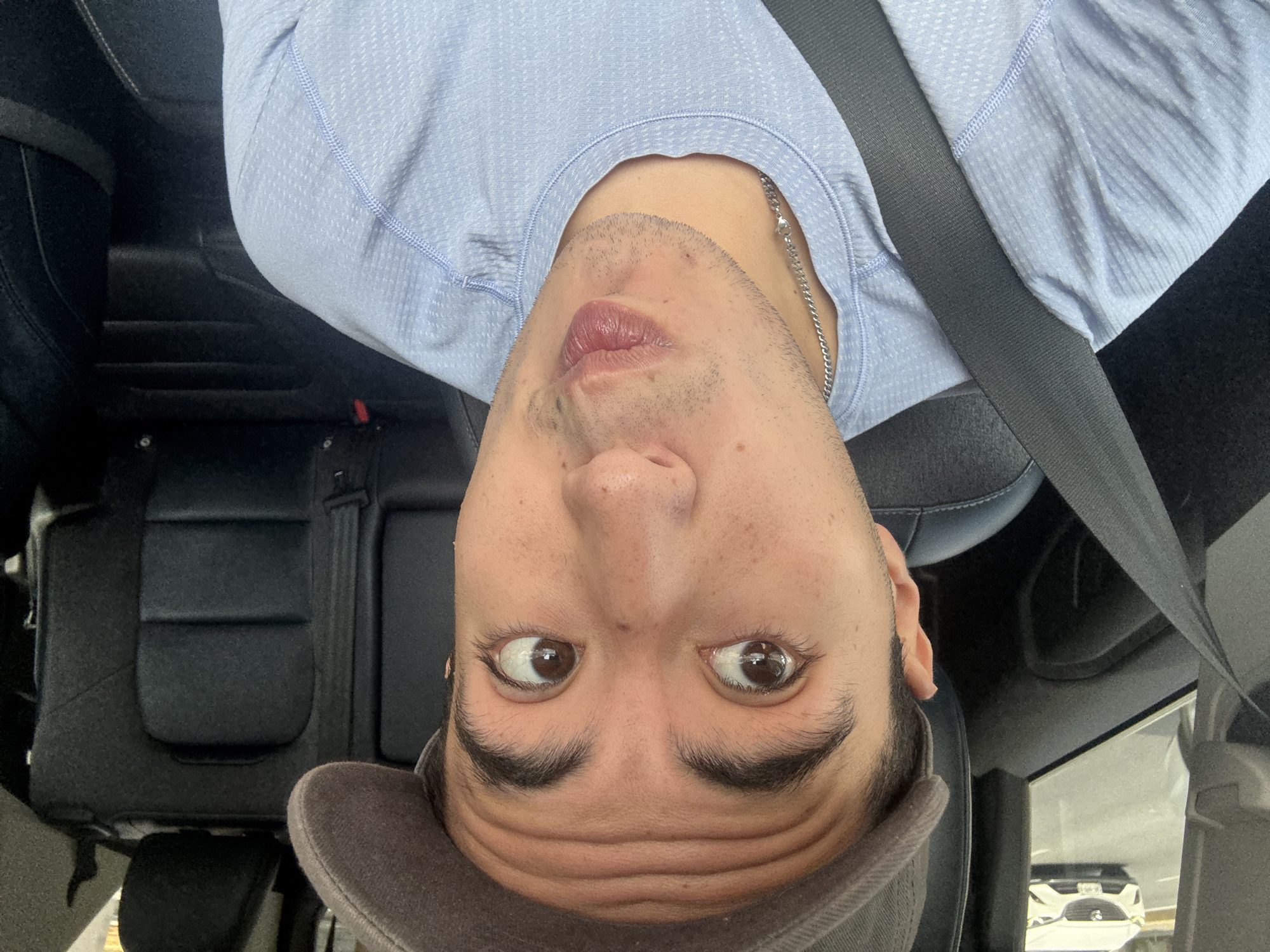 A medium-shot, front-facing selfie taken from inside a vehicle shows a person wearing a grey baseball cap and a light blue, textured t-shirt. The person is looking upwards and to their left, with their lips pursed. The background of the image reveals the interior of the car, including black leather seats and a seatbelt. Outside the vehicle, through the window, another white car is visible on a paved road. The person's face is the primary focus, with prominent eyebrows and freckles visible. A silver chain necklace is also apparent. The scene appears to be set in a city, with the presence of vehicles and infrastructure suggesting an urban environment. It is not possible to determine the specific location within Monterrey, Mexico based on the provided image.