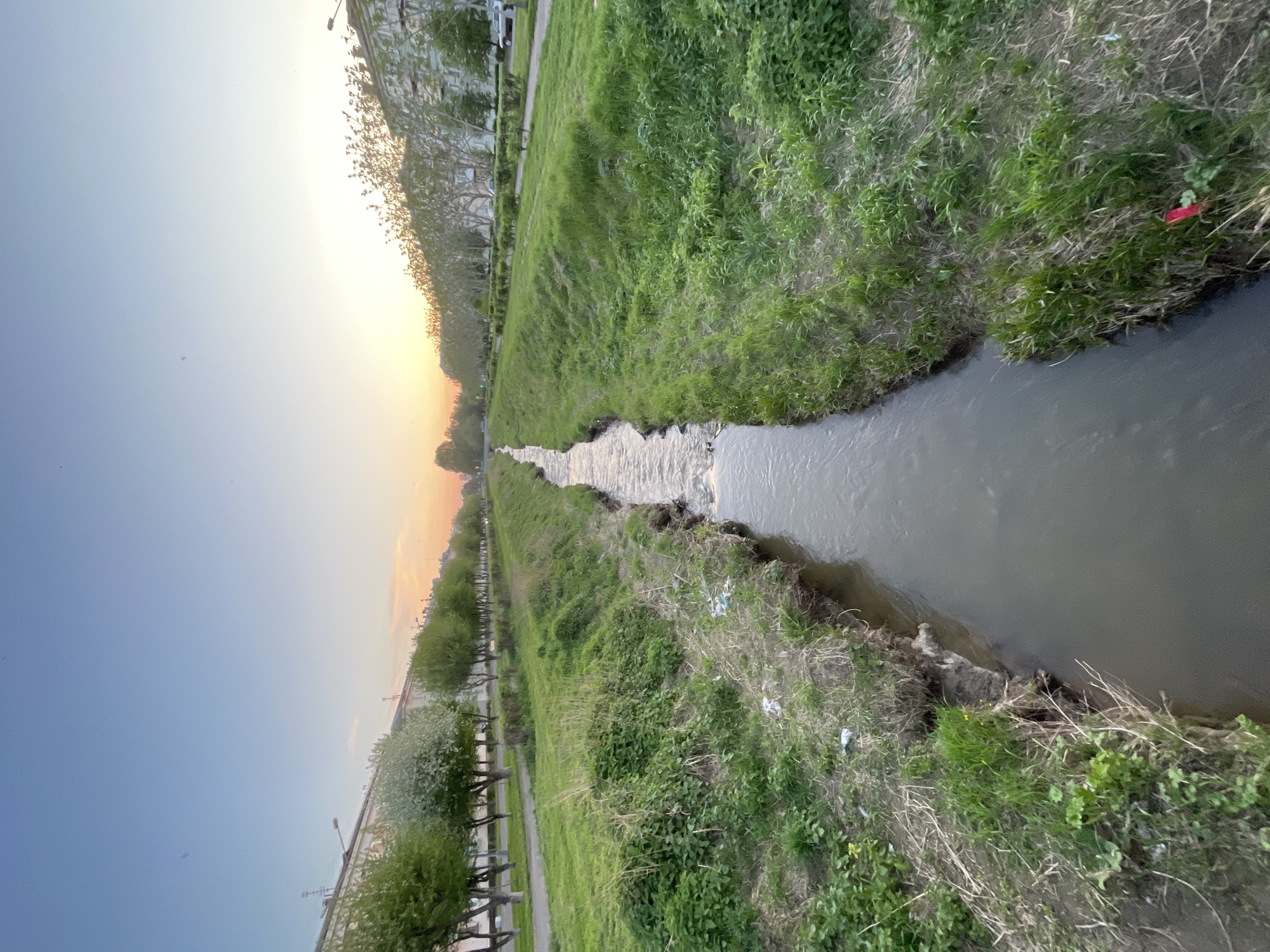The image depicts a narrow canal or waterway running through a grassy area. Buildings and trees line one side of the canal, while the opposite side is primarily grass and vegetation. The water appears murky. The sky is visible in the background with colors suggesting either sunrise or sunset. The image orientation is vertical, making the canal seem to run up and down. The overall scene suggests an urban or suburban area with a focus on the waterway. The location of the canal within Zheleznogorsk, Russia, cannot be determined solely from the image.