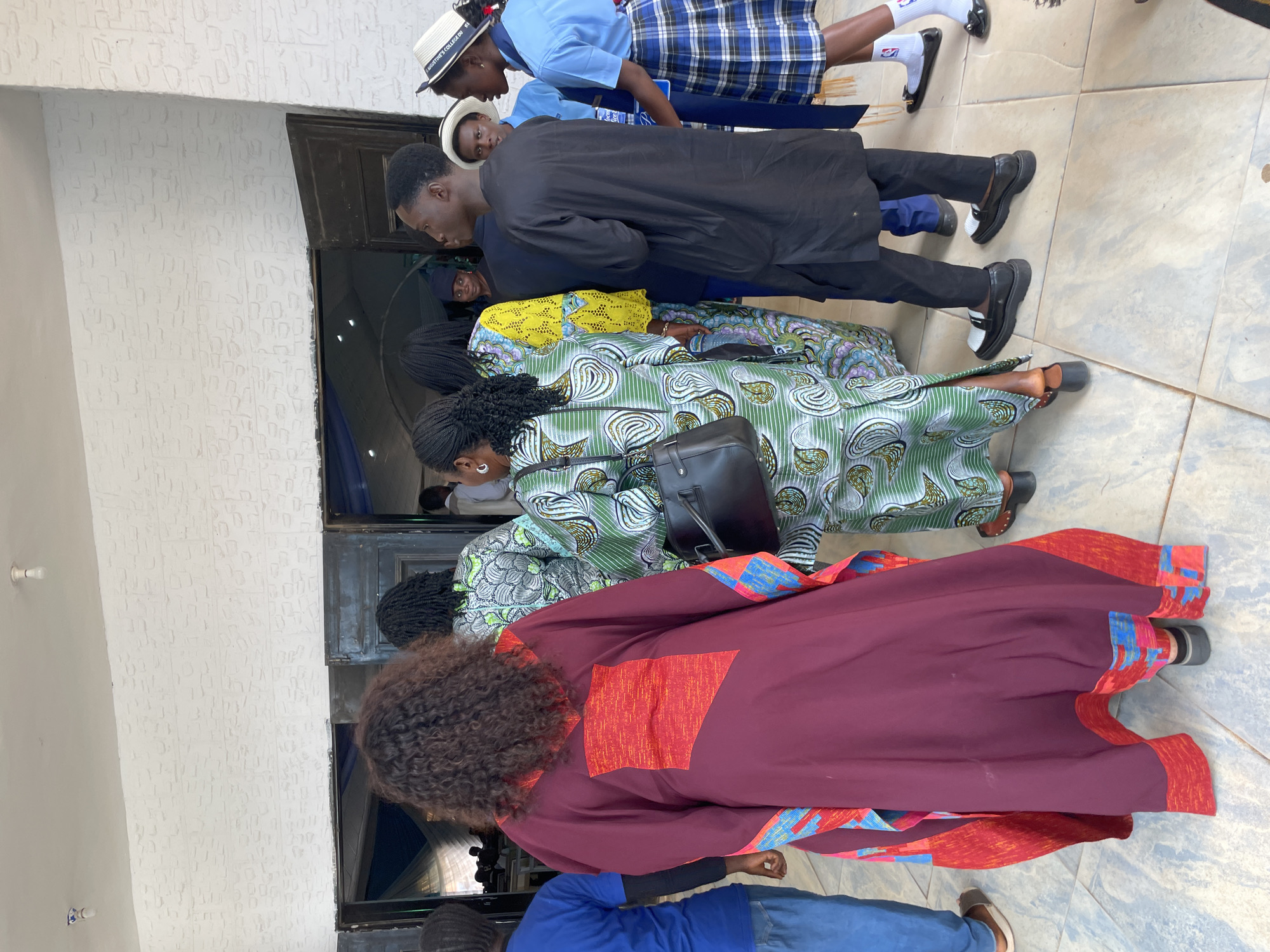 The image captures a gathering of people outside a building, possibly a school or community center, in Bar Gada, Nigeria. The architecture suggests a modern, possibly institutional setting, with a light-colored, textured wall and dark-framed doorways. A group of individuals, predominantly women and some young people, are seen from behind, facing away from the camera and towards an entrance.
Several people are dressed in colorful, patterned traditional attire, typical of Nigerian fashion. One woman in the foreground wears a striking, flowing maroon robe with vibrant red and blue geometric patterns on the sleeves and hem. Another woman is clad in a green and white patterned dress. A third woman sports a bright yellow outfit with a distinctive pattern on the sleeves.
Among the younger individuals, two appear to be students in uniform. One, a girl, wears a blue collared shirt and a blue and white plaid skirt, her hair neatly braided and a white hat with a blue band perched on her head. Another young person, a boy, is dressed in a black traditional garment. A few other individuals are partially visible in the background, including a person in a blue t-shirt and jeans.
The ground is paved with light-colored square tiles. The overall scene suggests an event, perhaps a ceremony or a gathering, with people awaiting entry into the building or socializing outdoors. The lighting indicates it is daytime, possibly with overcast conditions, as there are no harsh shadows. The atmosphere appears calm and orderly, with individuals engaged in conversation or simply standing together.