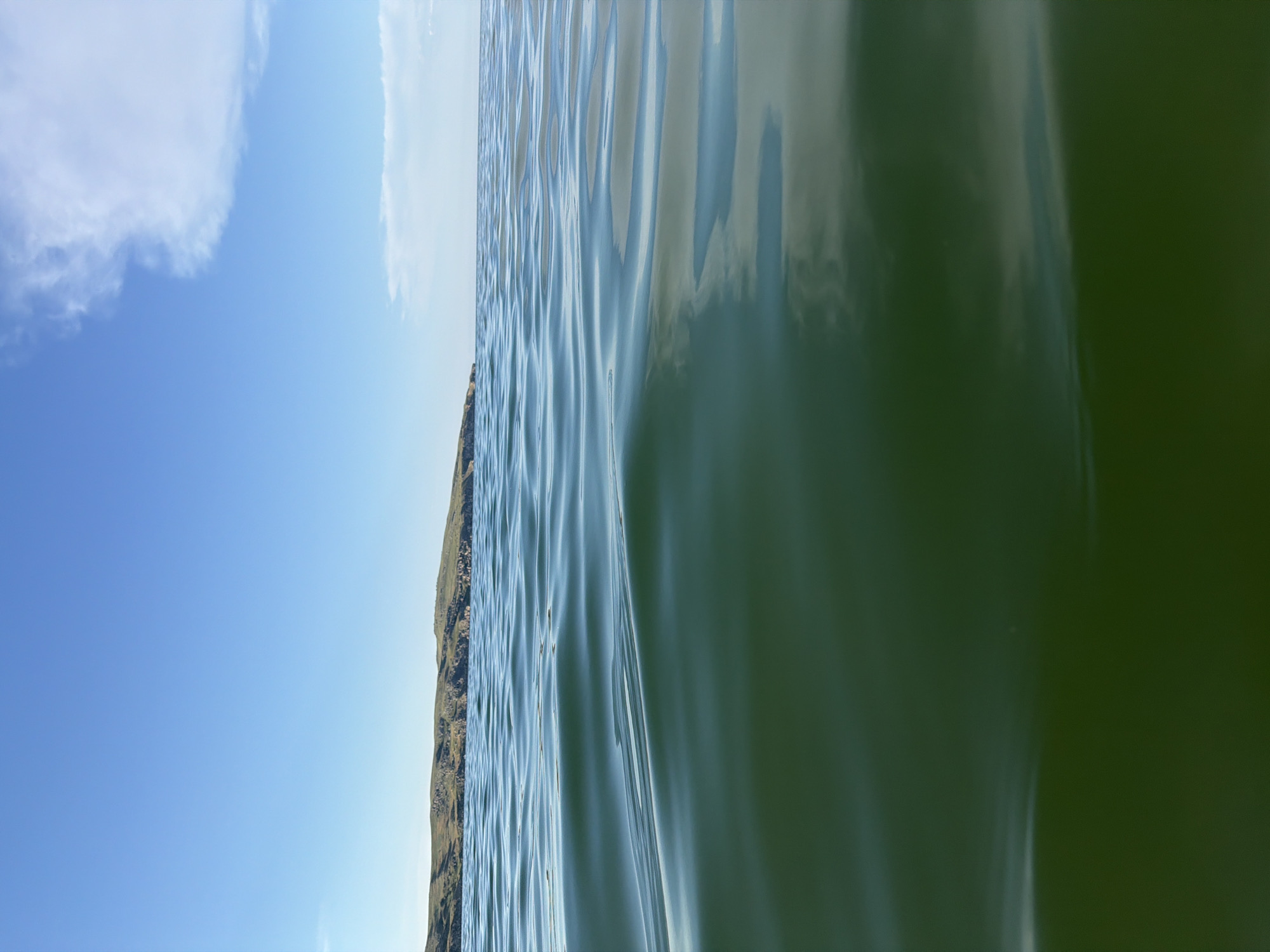 This is an eye-level, vertical, wide-angle shot of a calm, dark green body of water. The water fills the lower two-thirds of the frame, with subtle ripples on its surface reflecting the blue sky. In the upper third, a clear, bright blue sky transitions to a lighter hue towards the horizon. A thin strip of land with rolling hills and sparse vegetation is visible along the horizon line, separating the water from the sky. A few wispy white clouds are scattered across the sky. The image appears to be taken from a low vantage point, possibly from the surface of the water or a very low boat. The location is identified as Мысовское сельское поселение, Ukraine. There are no people or man-made objects clearly visible in the image.