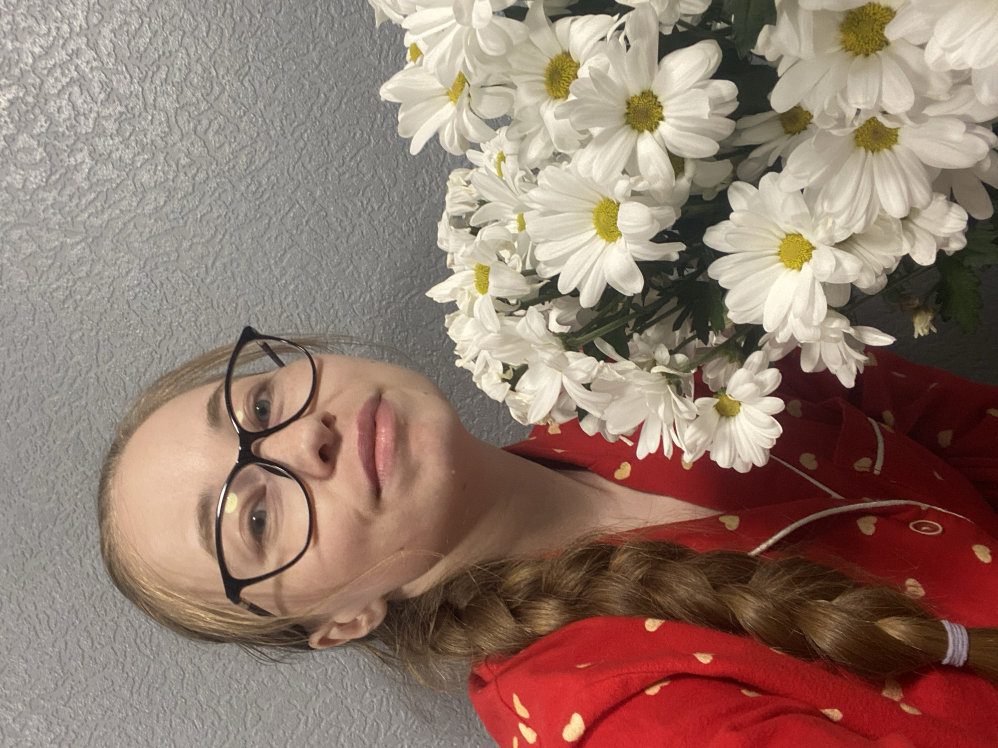 A woman with blonde hair braided and wearing black-rimmed glasses poses with a bouquet of white daisies with yellow centers. She is wearing a red pajama top with small, faint gold polka dots. The background is a textured gray wall. There is no evidence to suggest that this image was taken in Saint Petersburg, Russia.