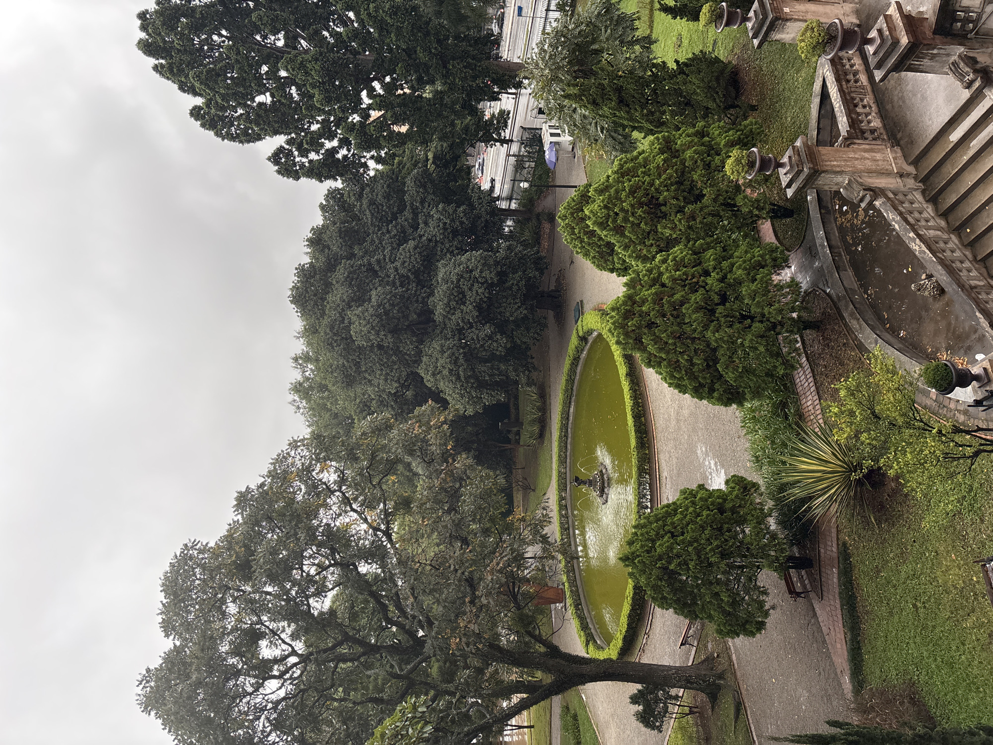 An elevated, slightly rotated view of a section of Parque da Independência in São Paulo, Brazil, under a cloudy sky. The park features a large oval fountain with water, enclosed by trimmed hedges, situated between walking paths. Mature trees surround the fountain, providing shade. A grand staircase with decorative stone balustrades borders the edge of the frame. The scene includes manicured lawns, decorative plants in urns atop the stone structures, and glimpses of a street with vehicles in the background. There are no people immediately visible in the photograph.