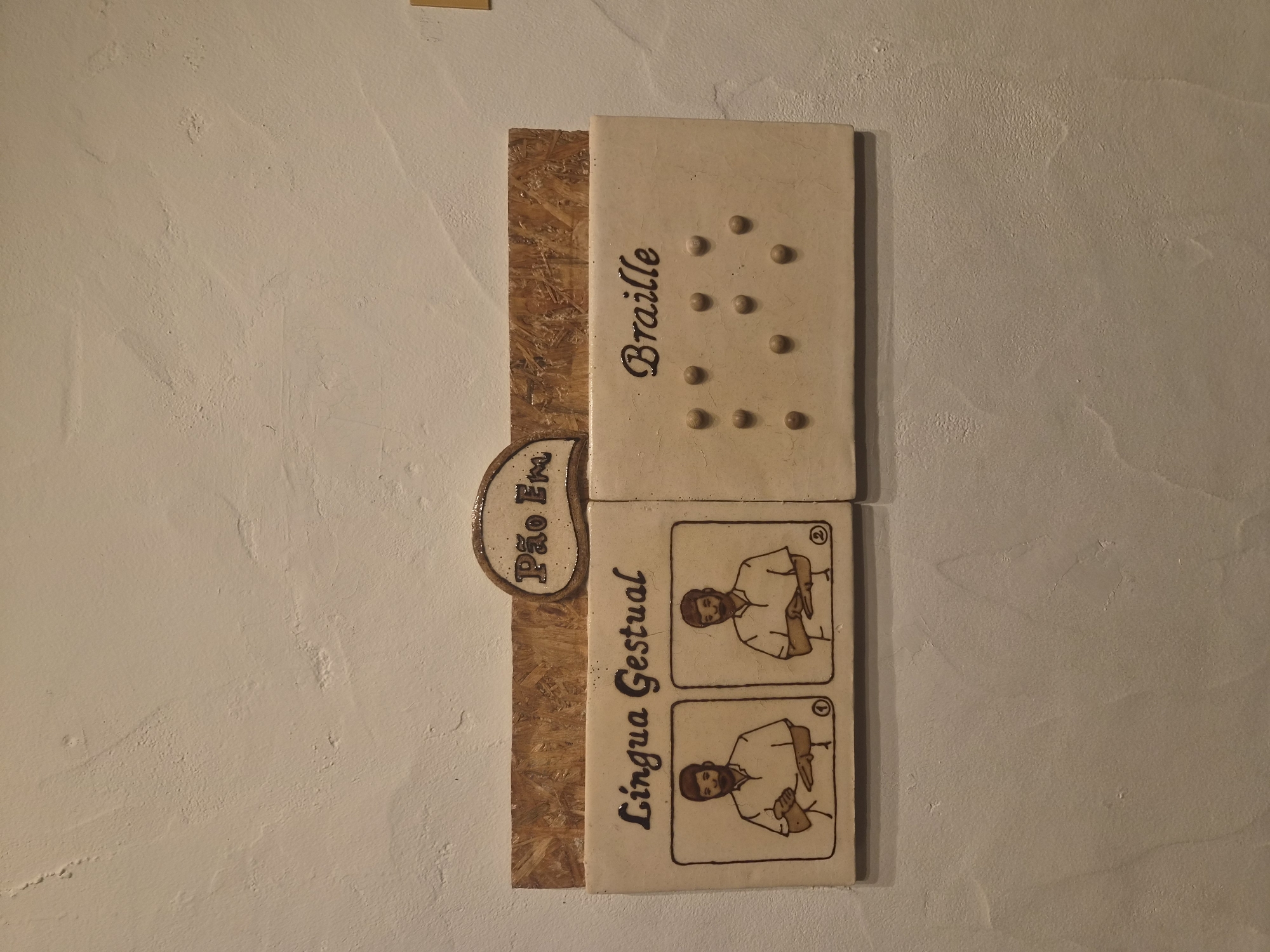 This image shows a sign on a textured, off-white wall. The sign is mounted on a piece of wood and has two sections. The left section, labeled "Lingua Gestual" (Gestural Language), features two framed illustrations of a person making hand gestures. The right section, labeled "Braille", displays a pattern of raised dots, characteristic of Braille writing. Above these sections, a small, oval-shaped sign reads "Pão Em" (Bread In). Based on its presence in Seia, Portugal, this sign likely serves as an informational or accessibility feature, possibly indicating that bread is available or that information about bread is provided in both gestural language and Braille.