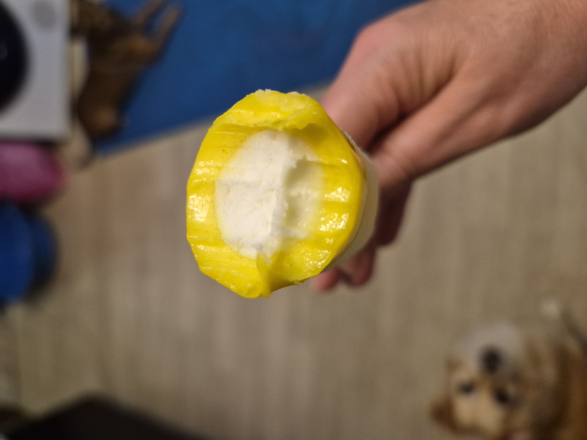An extreme close-up shot shows a person's hand holding a partially eaten yellow popsicle, with the white ice cream interior visible. The background is blurred, showing a tan floor, a blue mat, and out-of-focus objects including what appears to be a dog. The scene is indoors, and there is no information to suggest this is in Nyagan, Russia.