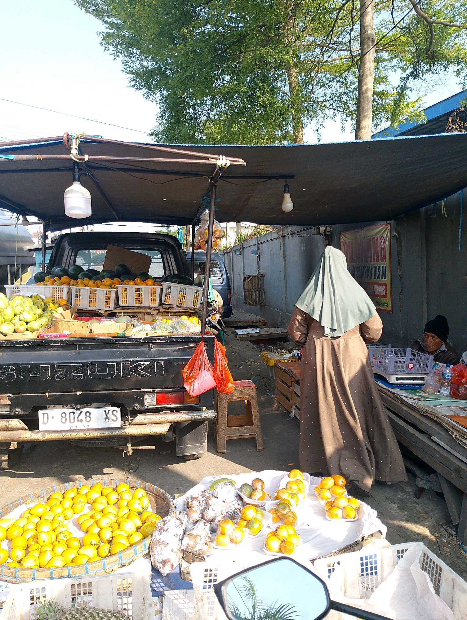 A fruit stand is set up on the side of a street in Lembang, Indonesia. A black pickup truck, possibly a Suzuki, is loaded with various fruits including watermelons, oranges, and other produce in white crates. A woman wearing a light green hijab and a long brown dress stands facing away from the camera, looking at the fruit. To the right, a man wearing a black beanie sits behind a table with a scale and plastic bags of produce. The scene is outdoors, with trees and a building in the background. The ground is paved, and there are some debris scattered around. A motorcycle's side mirror is visible in the foreground, reflecting palm fronds.