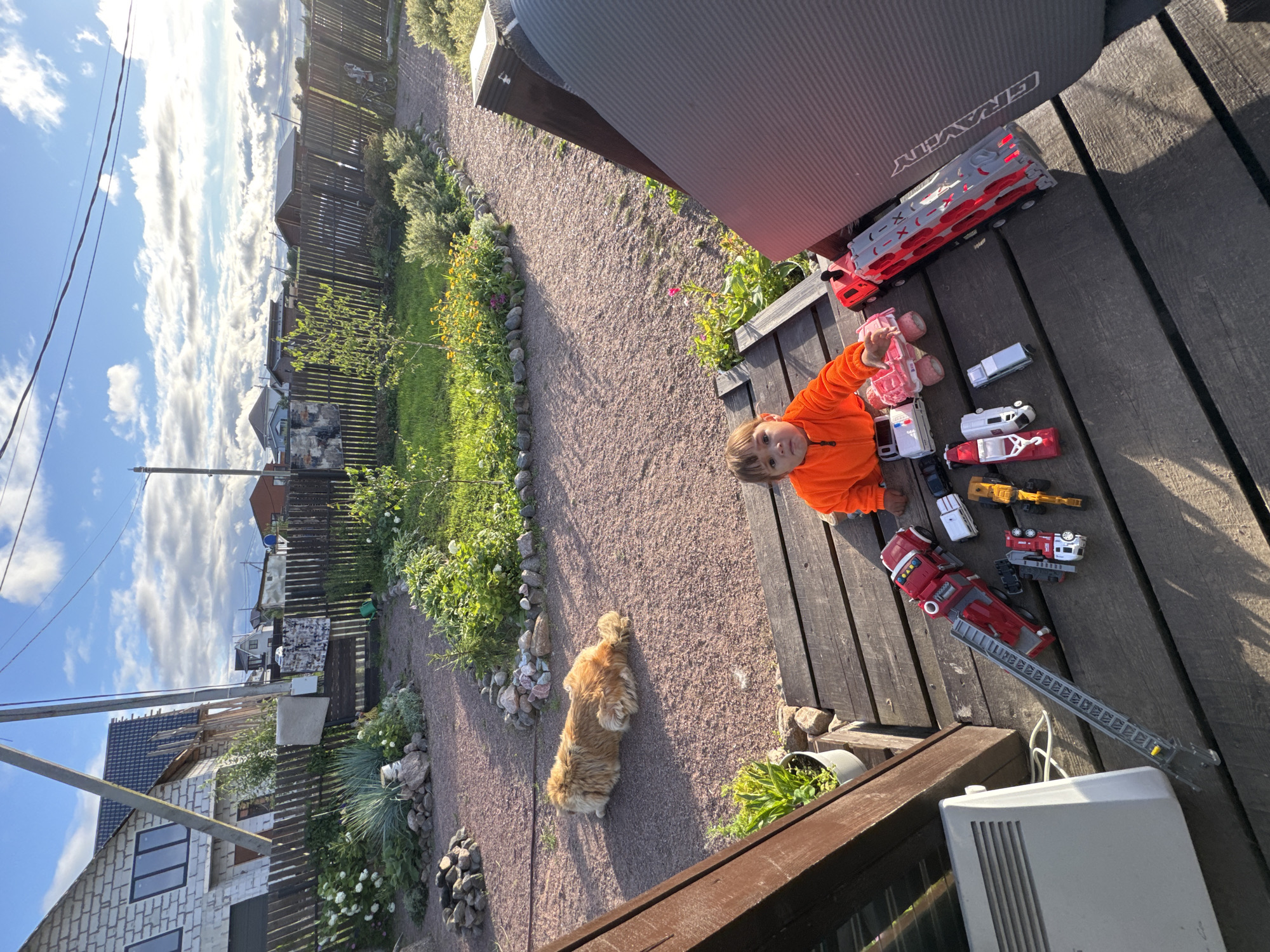 In a backyard setting in Kempelevo, Russia, a young child wearing an orange jacket sits on wooden planks, surrounded by toy vehicles. The child is looking directly at the camera. Among the toys are a red fire truck with an extendable ladder, a white ambulance, a pink truck, and other assorted toy vehicles. To the left of the child, a white radiator is visible. In the background, a wooden fence encloses a garden with green grass, flower beds, and a paved path. A fluffy, light-colored dog is lying on the gravel path in the middle ground. The scene is bathed in the warm light of what appears to be late afternoon or early evening.