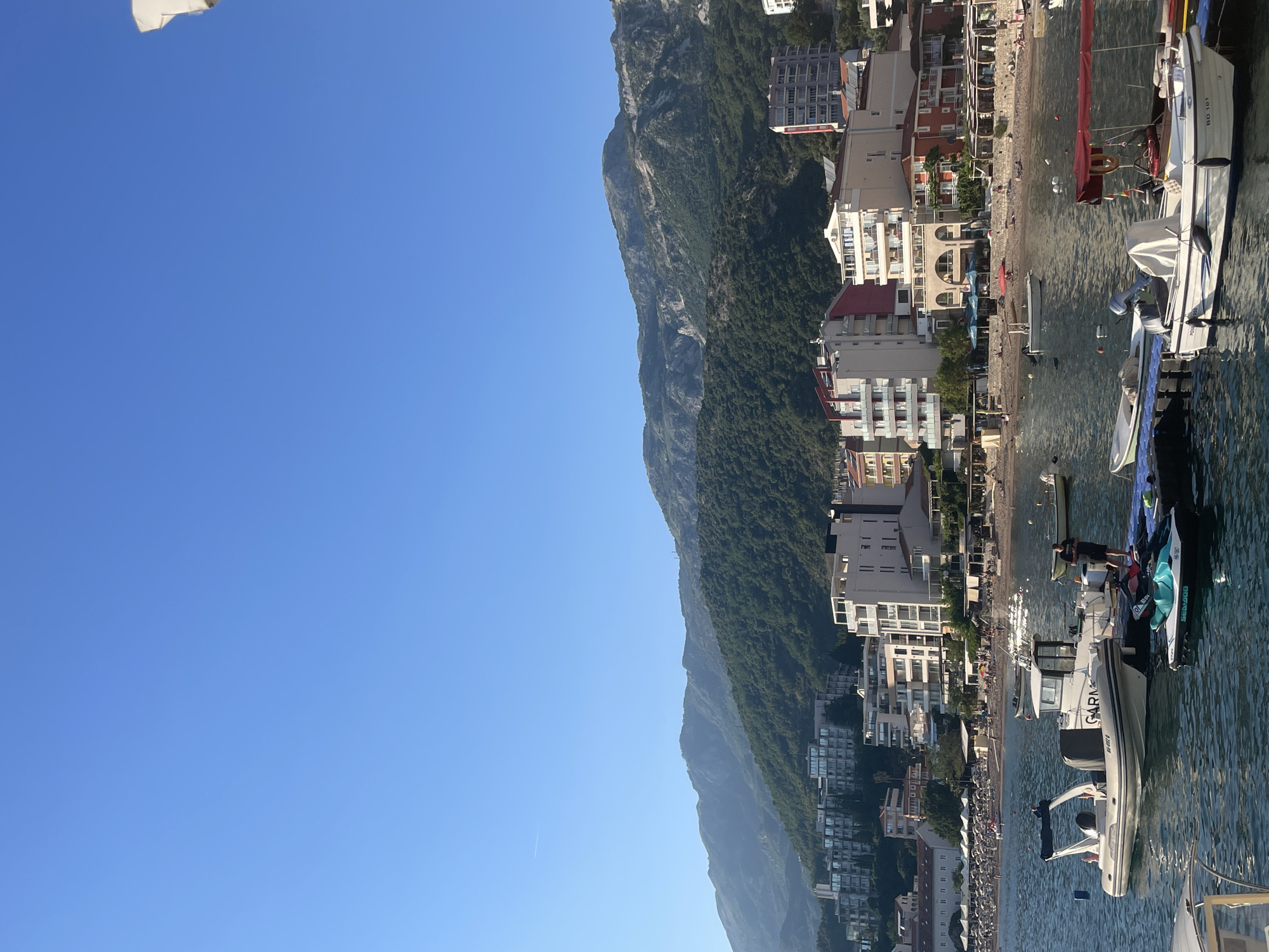 The image captures a coastal scene in Bečići, Montenegro, featuring a combination of natural and urban elements. The sky is a clear, gradient blue, taking up a significant portion of the frame. A towering, green, densely vegetated mountain range forms a vertical barrier along the right, extending into the background. The town of Bečići is built at the base of the mountains, visible as a series of buildings with varying heights and architectural styles. These structures include both residential and commercial properties.
The shoreline contains a sandy beach crowded with people. The beach transitions into the turquoise water of the Adriatic Sea, where several boats are moored. The boats in the foreground, near a pier, range from small vessels to larger yachts. A speedboat labelled with "GARMIN" is prominent near the bottom right of the frame.