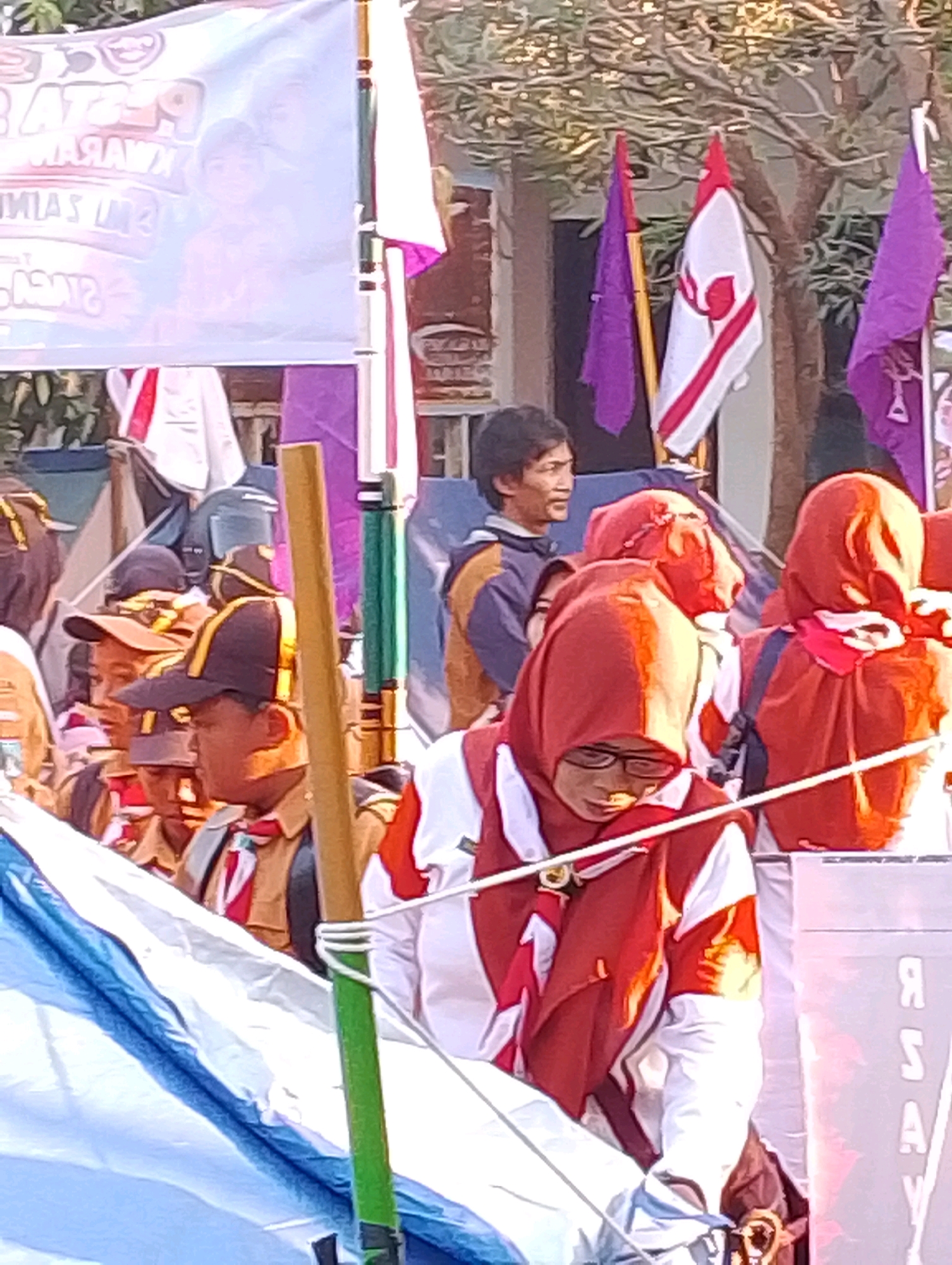 The image depicts a group of people, likely scouts, at an outdoor event in Probolinggo, Indonesia. In the foreground, a person wearing a red headscarf and glasses is visible, with a blue and white tent or banner in front of them. Behind this person, other individuals in scout uniforms with brown and yellow sashes are present. Several flags, including purple and white ones with red accents, are displayed in the background. A man with dark hair is looking to the right. The setting appears to be a street or open area, possibly a park or school grounds, with trees and buildings visible in the distance. The overall atmosphere suggests a community gathering or event.