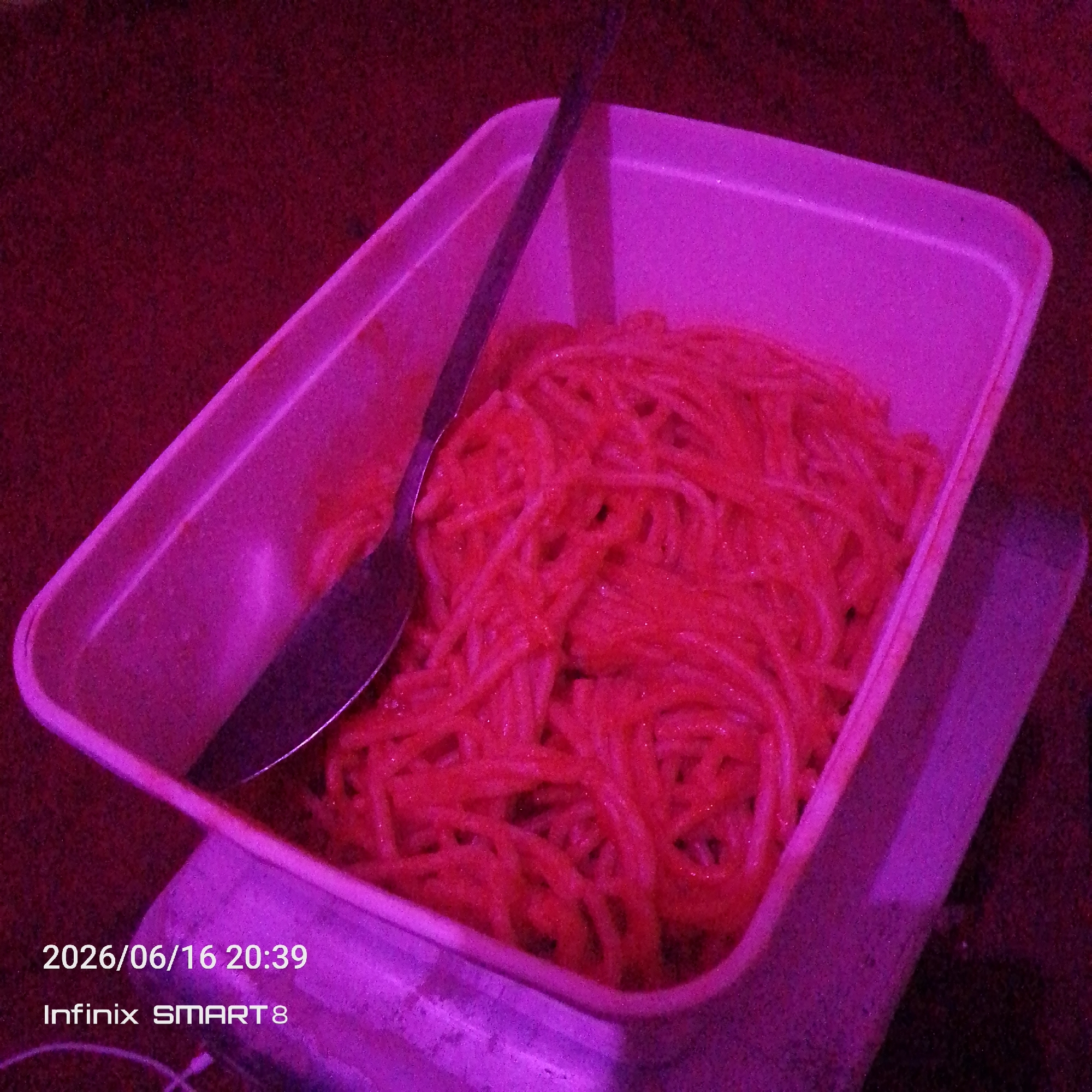 The image shows a white plastic container filled with spaghetti coated in a red sauce, with a metal spoon resting on top. The lighting is a vivid purple, casting a strong color cast over the entire scene. The container and food are positioned on a textured, dark red surface. In the bottom left corner, a timestamp and device name are visible, reading "2026/06/16 20:39" and "Infinix SMART 8". There are no people or any discernible elements indicating a specific location within the city of Jos, Nigeria.