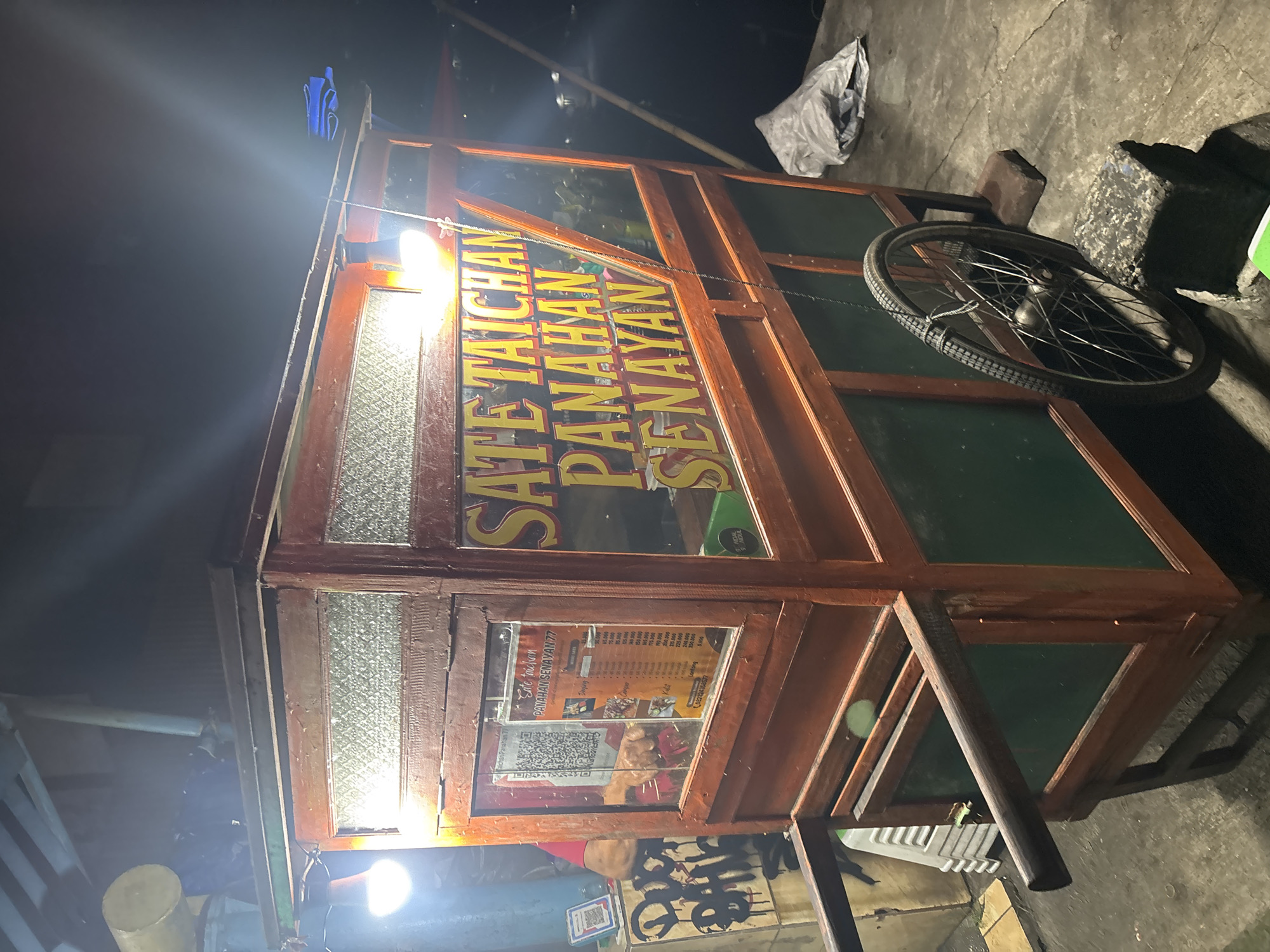 This vertical image displays a street food cart, identified as "SATE TAICHAN PANAHAN SENAYAN," operating at night in Bogor, Indonesia. The wooden cart features glass panels, some clear and some textured, and is brightly illuminated by an artificial light source, creating lens flares and reflections.
The main subject is the food cart, with its prominent yellow signage clearly visible. Inside one of the cart's compartments, a menu is displayed, listing various items such as "Daging" (meat), "Kulit" (skin), and "Lontong" (rice cake), along with prices like 140.000, 90.000, and 250.000. A QR code for payment is also visible on the menu. A person's arm, wearing a red sleeve, is faintly discernible behind the menu, indicating a vendor is present or recently was.
The scene is set outdoors on a concrete or paved surface, characterized by the darkness of night contrasted with the bright light from the stall. A bicycle wheel rests against the right side of the cart, alongside a white plastic bag and a dark block. The overall ambiance suggests a busy, late-night food selling operation. The name "Panahan Senayan" points to a famous satay style originating from Jakarta, now likely sold in Bogor.