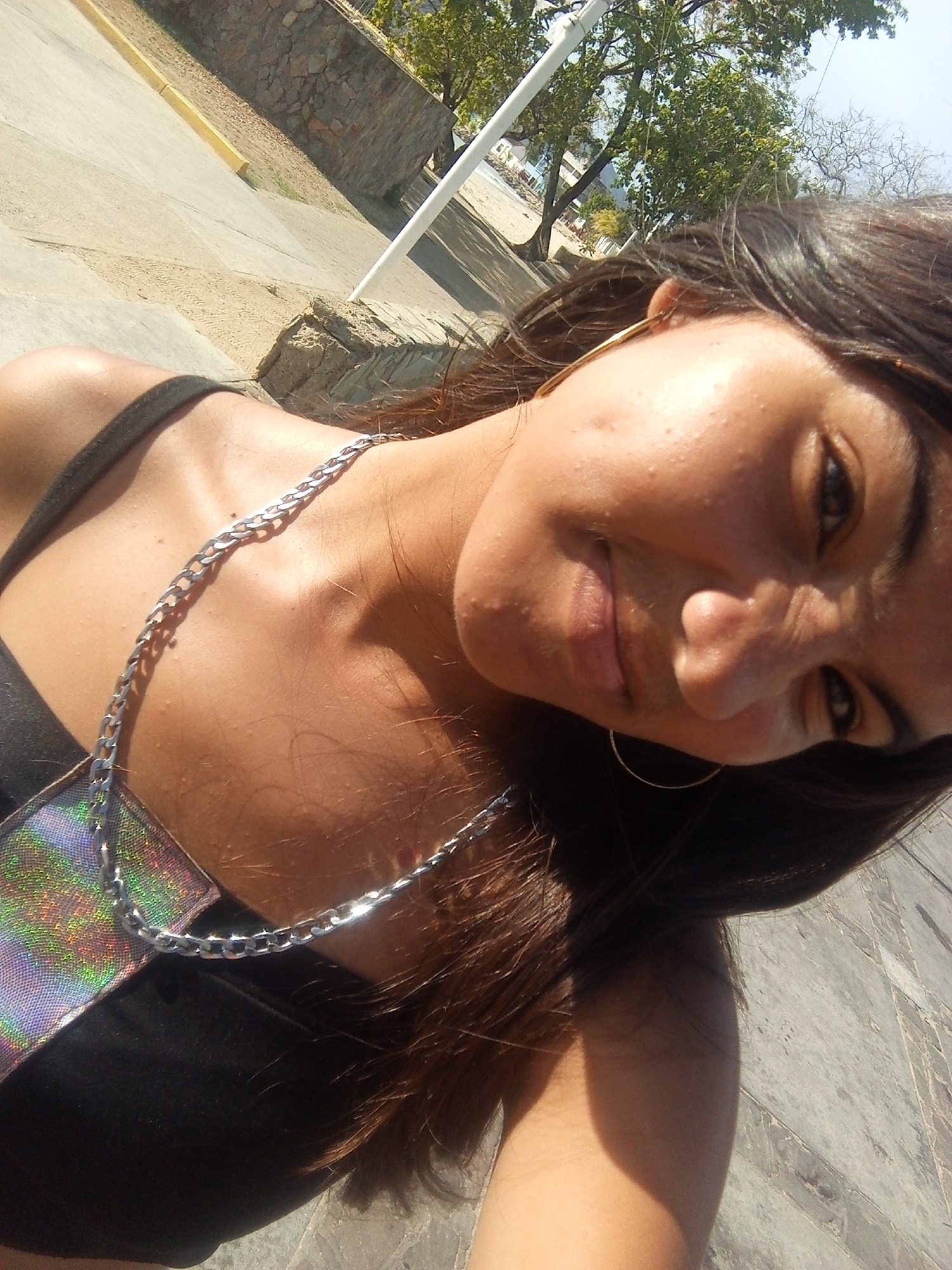 A young woman is captured in a tilted, close-up outdoor selfie in Juan Griego, Venezuela, under bright daylight. She has long, dark hair and looks towards the camera with a subtle smile. Her face is illuminated by harsh, direct sunlight, creating distinct shadows. She is wearing a black top with a prominent silver chain-link strap over her left shoulder, and a piece of iridescent or holographic fabric is visible near her chest, reflecting various colors. A golden hoop earring adorns her left ear.
The background, viewed from an elevated perspective, shows a paved ground that transitions into sandy or dusty patches. A low stone wall is visible, along with green trees under a clear, bright sky, indicating a sunny day. A white pole stands vertically in the mid-ground. Hints of a beach and distant buildings can be discerned through the foliage, consistent with a coastal town. No visible text is present in the image.